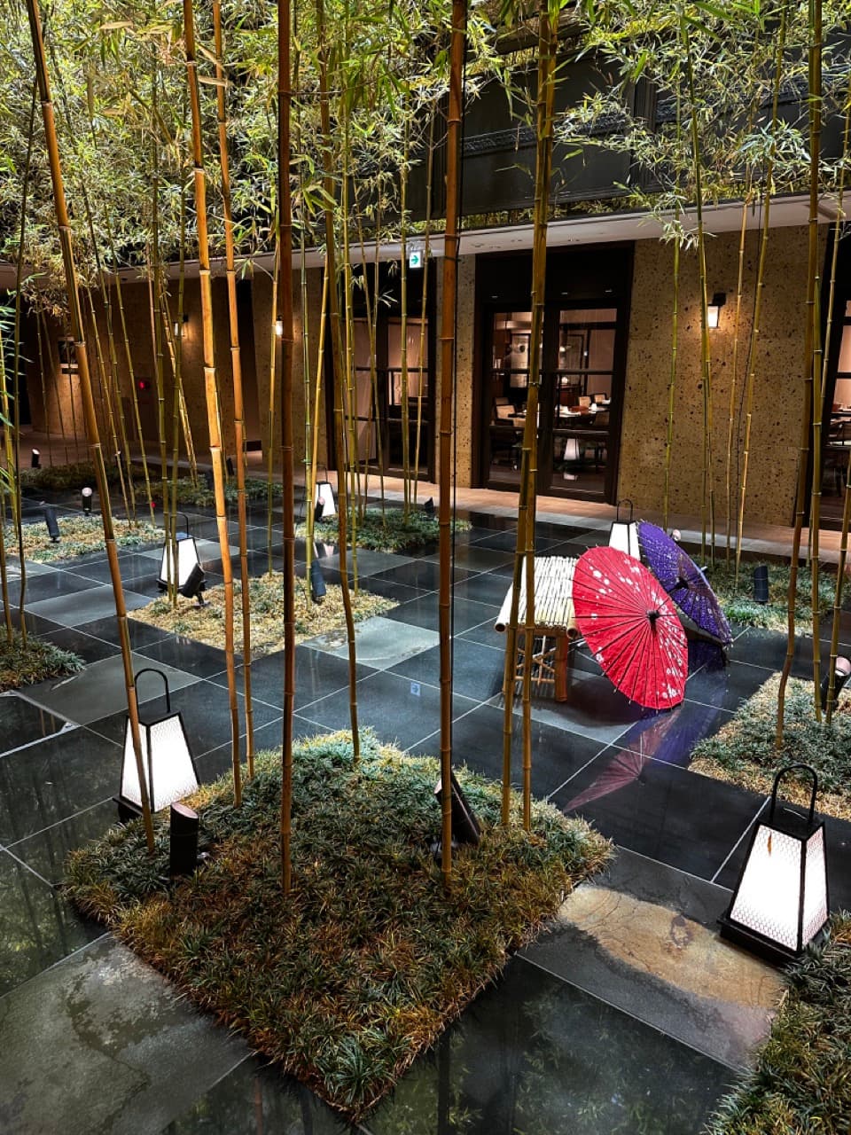 Bamboo growing in square patches with a black tile floor connecting each patch and a bamboo bench with a red and purple umbrella next to it, outside a small bamboo building with glass doors at Dhawa-Yura-Kyoto-Bamboo-Garden.