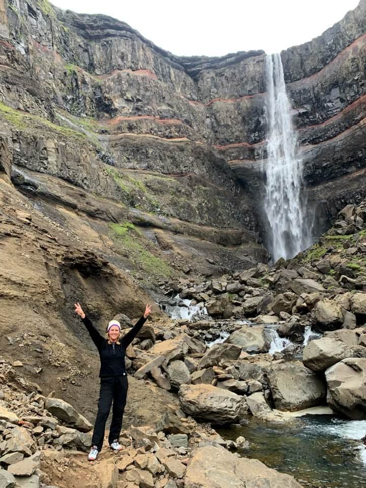 A person posing for a photograph with their arms in the air in front of a cliffside waterfall