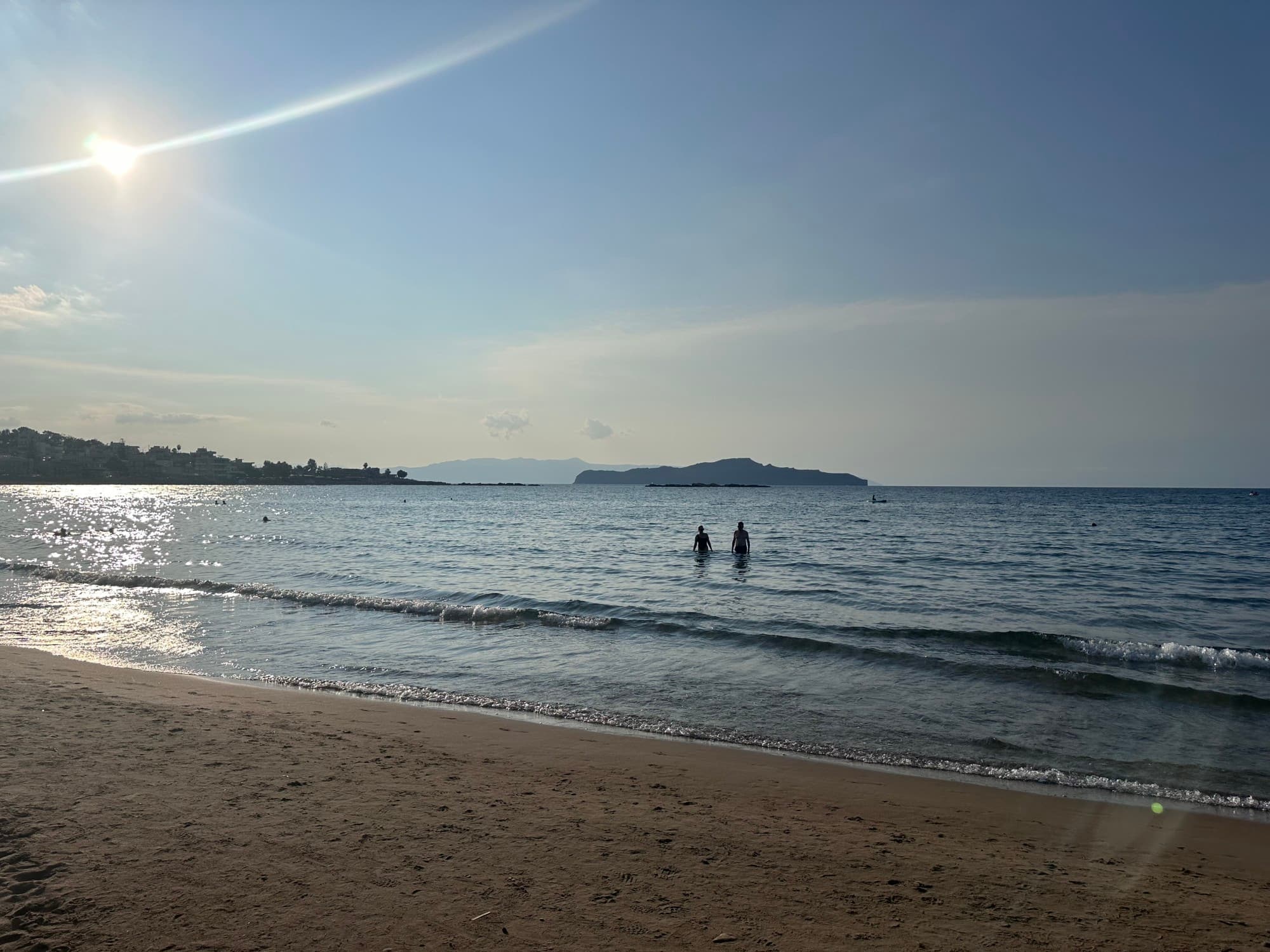 The sun and a sun flare over the ocean as two people wade into small waves off the beach.