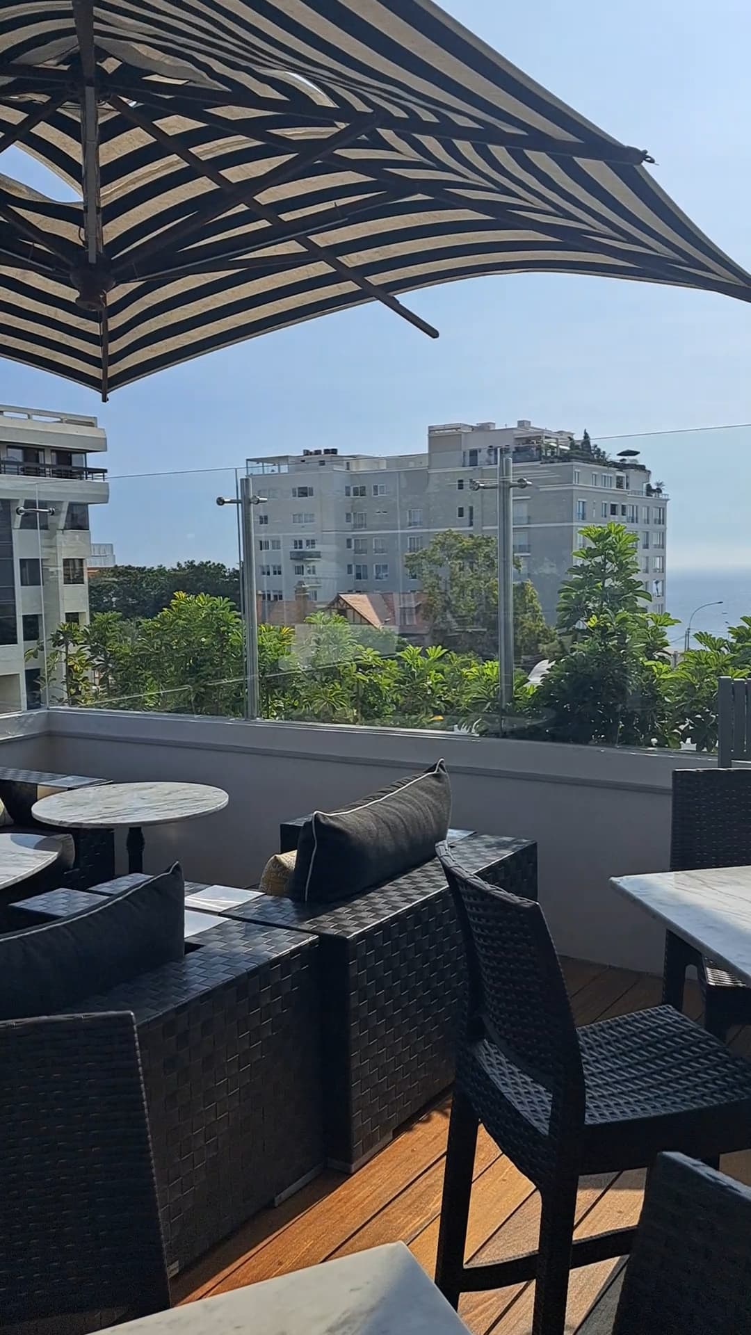 A view of the rooftop with black chairs and cabana seating underneath a black and cream striped umbrella. There are buildings and trees in the distance.