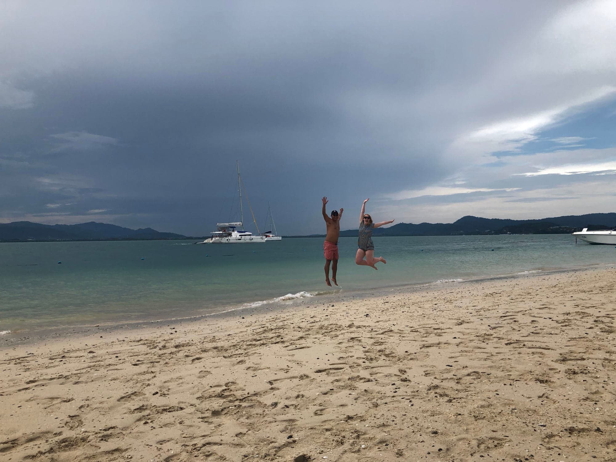 Couple jumping on the beach with a boat in the background and stormy clouds overhead.