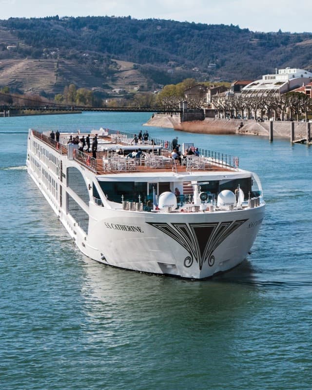 A large, white cruise ship sailing along a river with mountains in the distance