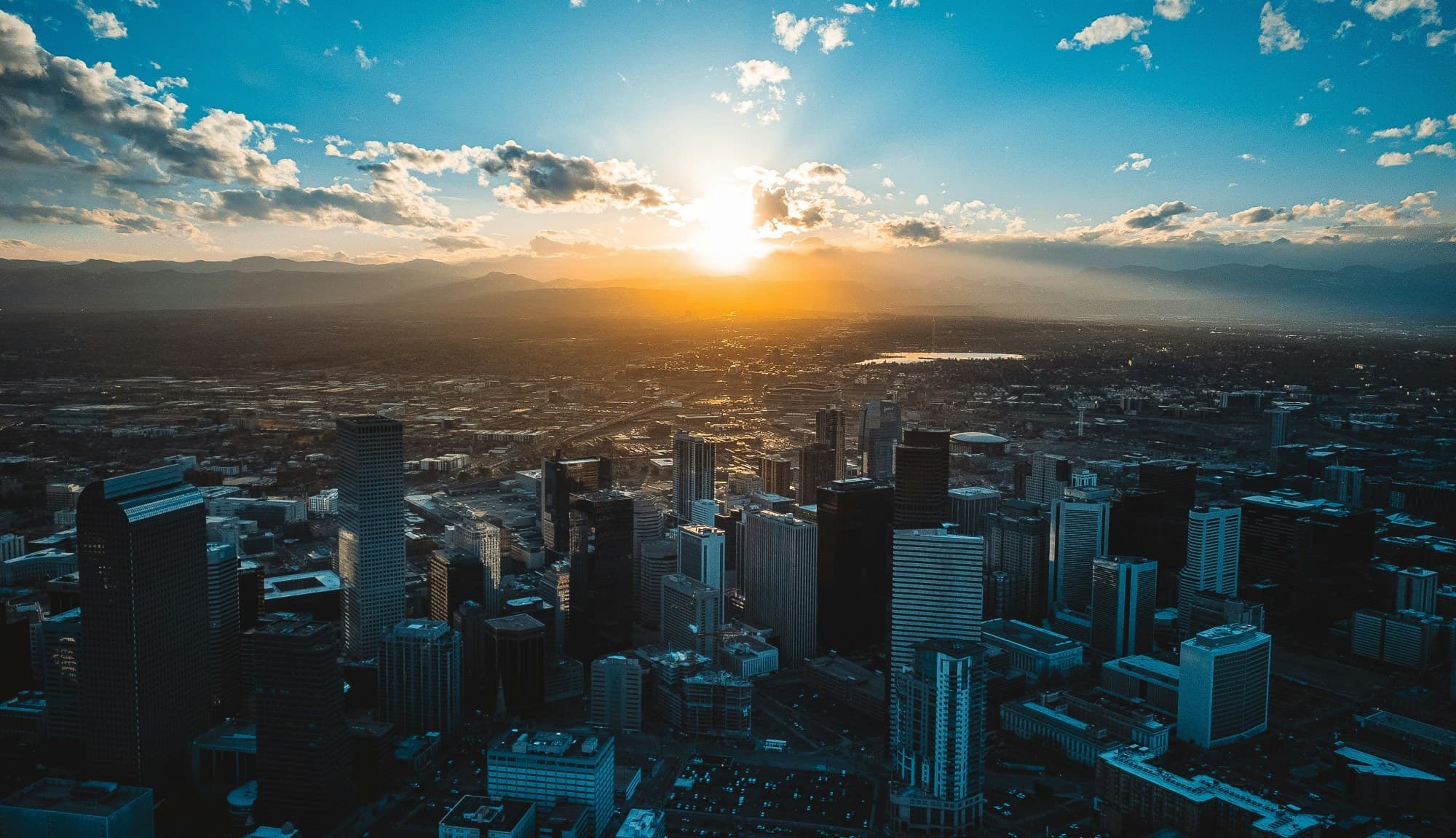 A bird's eye view of the city of Denver as the sun sets over the mountains with clouds reaching toward the horizon.