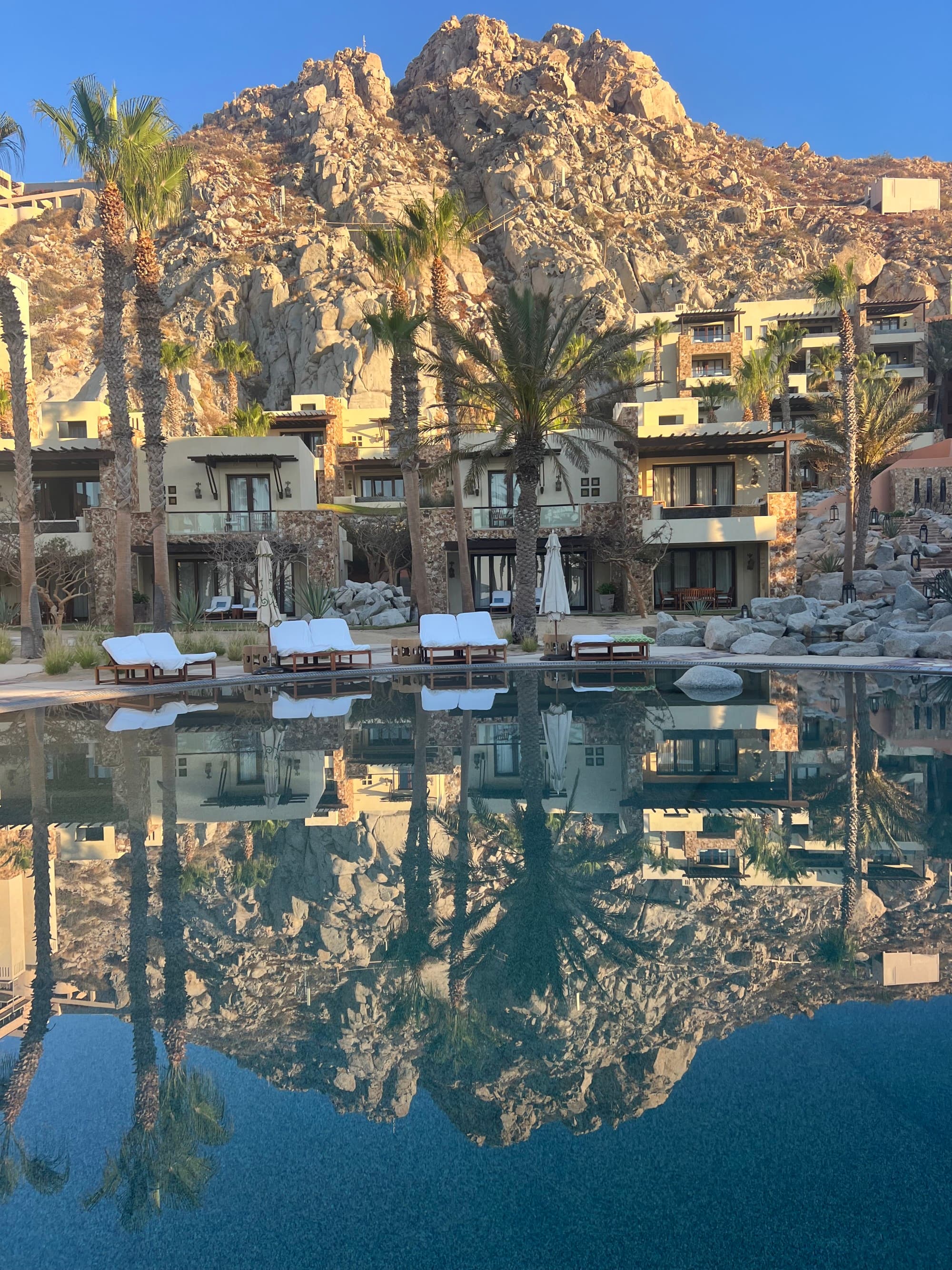 A serene pool area at a resort with palm trees and mountains in the background and their reflection in the water