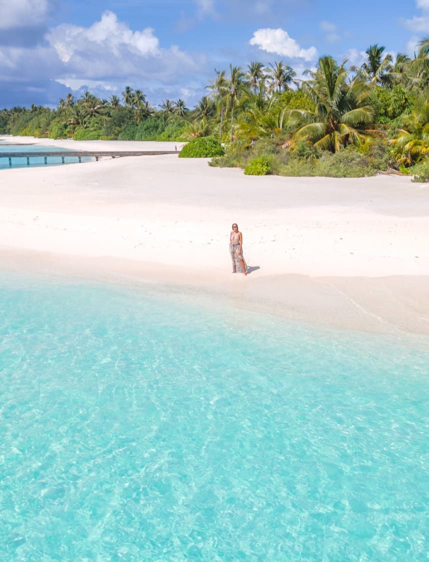 A person stands on a white sandy beach with bright blue ocean water in front and a tropical forest in the background.
