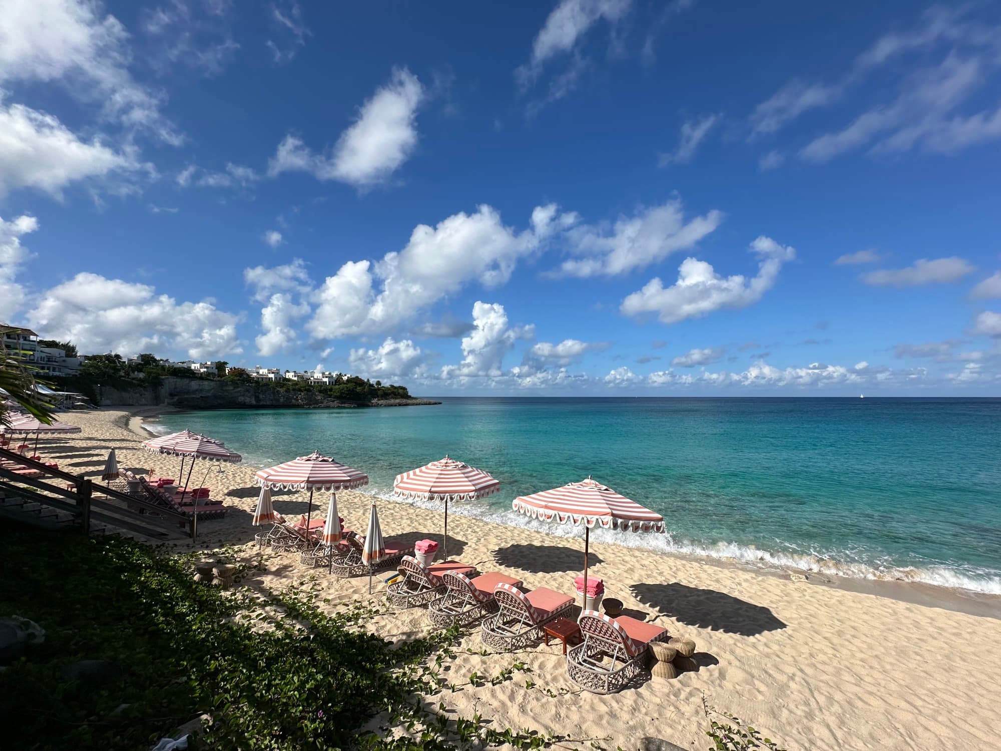 Loungers and umbrellas line the shore as gentle waves lap the shore on a sunny day dotted with clouds.