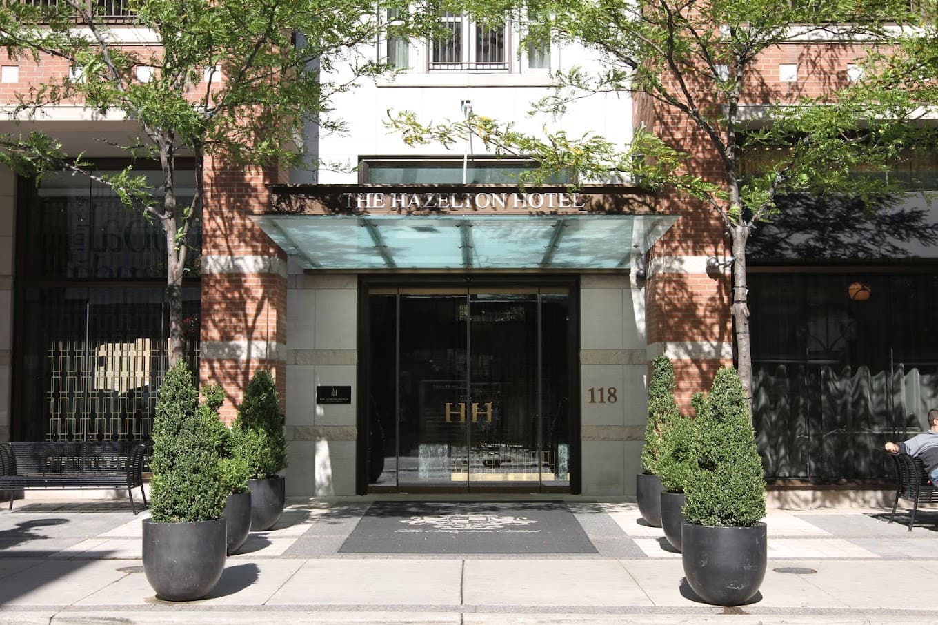 A picture of the elegant street entrance of the Hazelton Hotel, Toronto with plant pots either side of the entrance and glass doors and green and white exterior.
