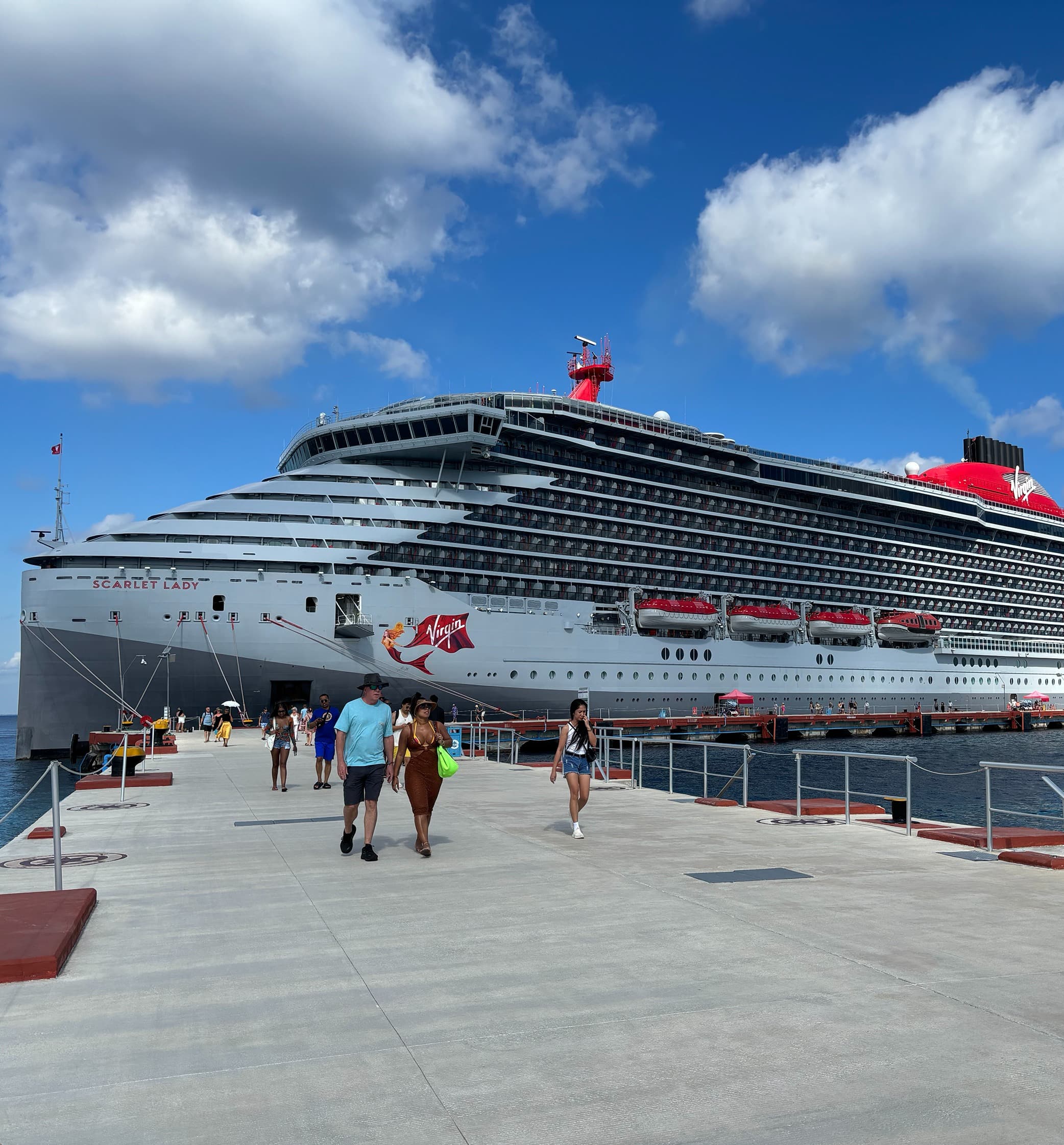 Tourists walking on a concrete pier away from the Virgin Voyages' Scarlet Lady cruise ship.
