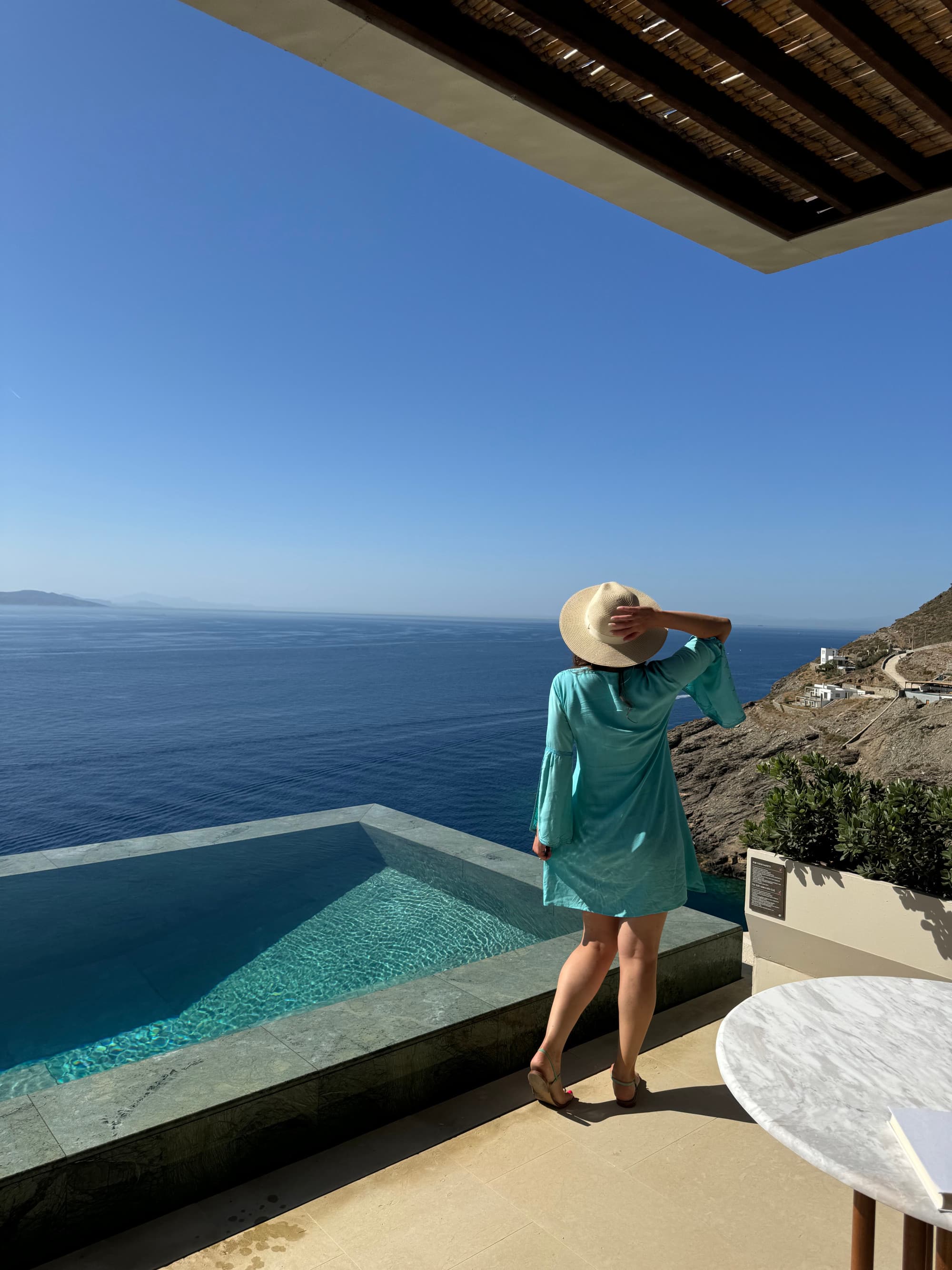 Eliza in a blue dress and sun hat standing by an infinity pool overlooking the ocean.
