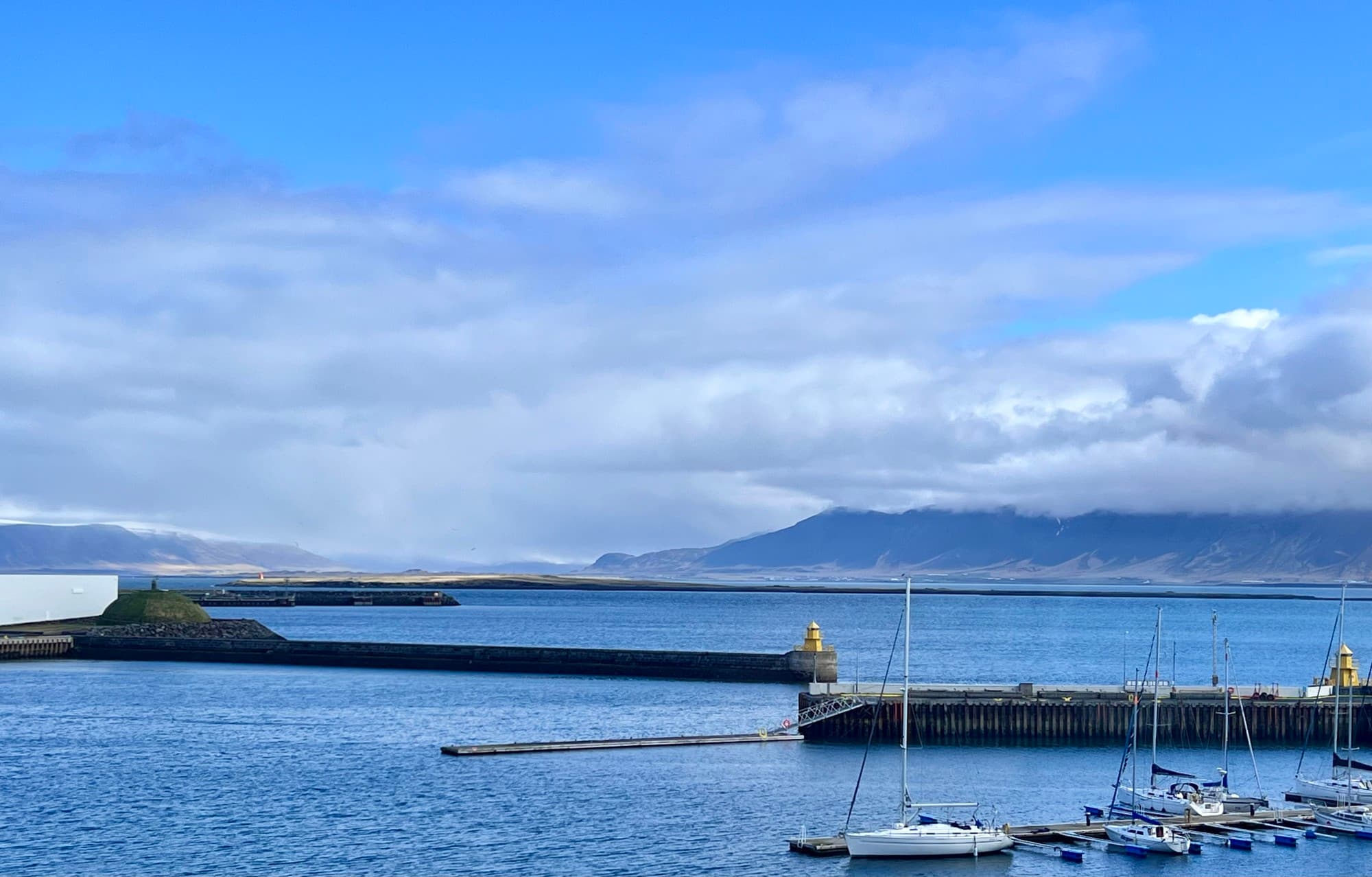 A serene view of the ocean with boats docked, a pier and moutains in the background with clouds overhead.