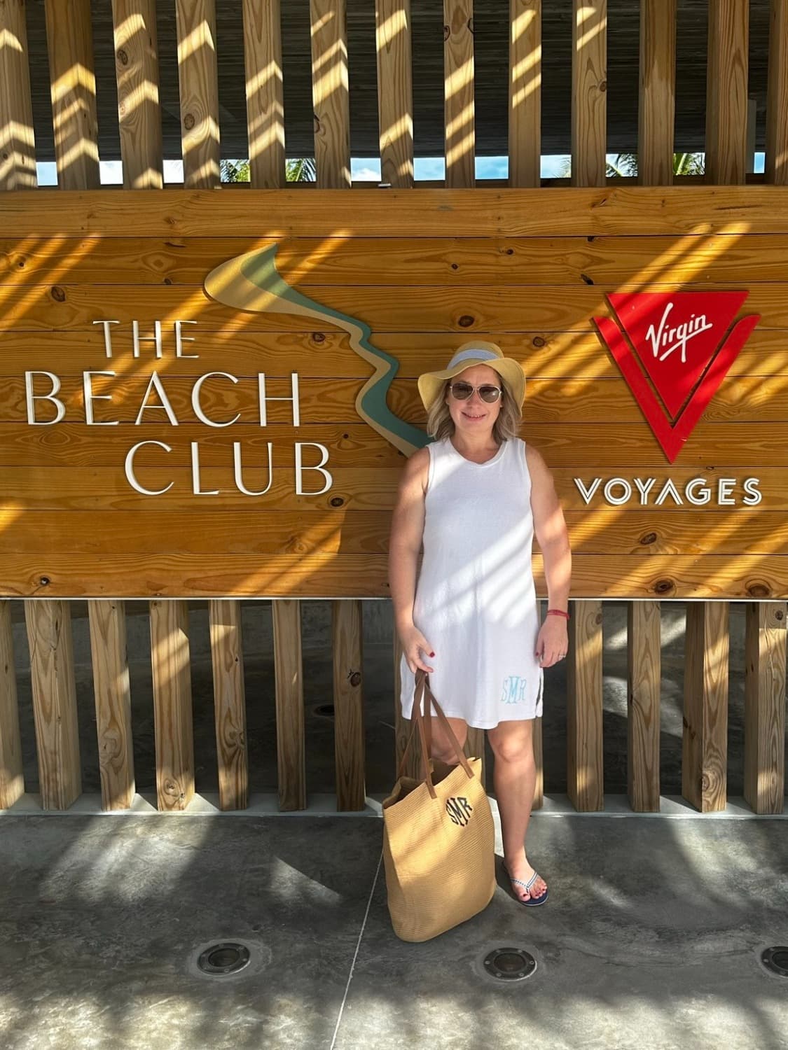Shelly Maffia in a white outfit and sun hat standing in front of a wooden sign that says "The Beach Club - Virgin Voyages"