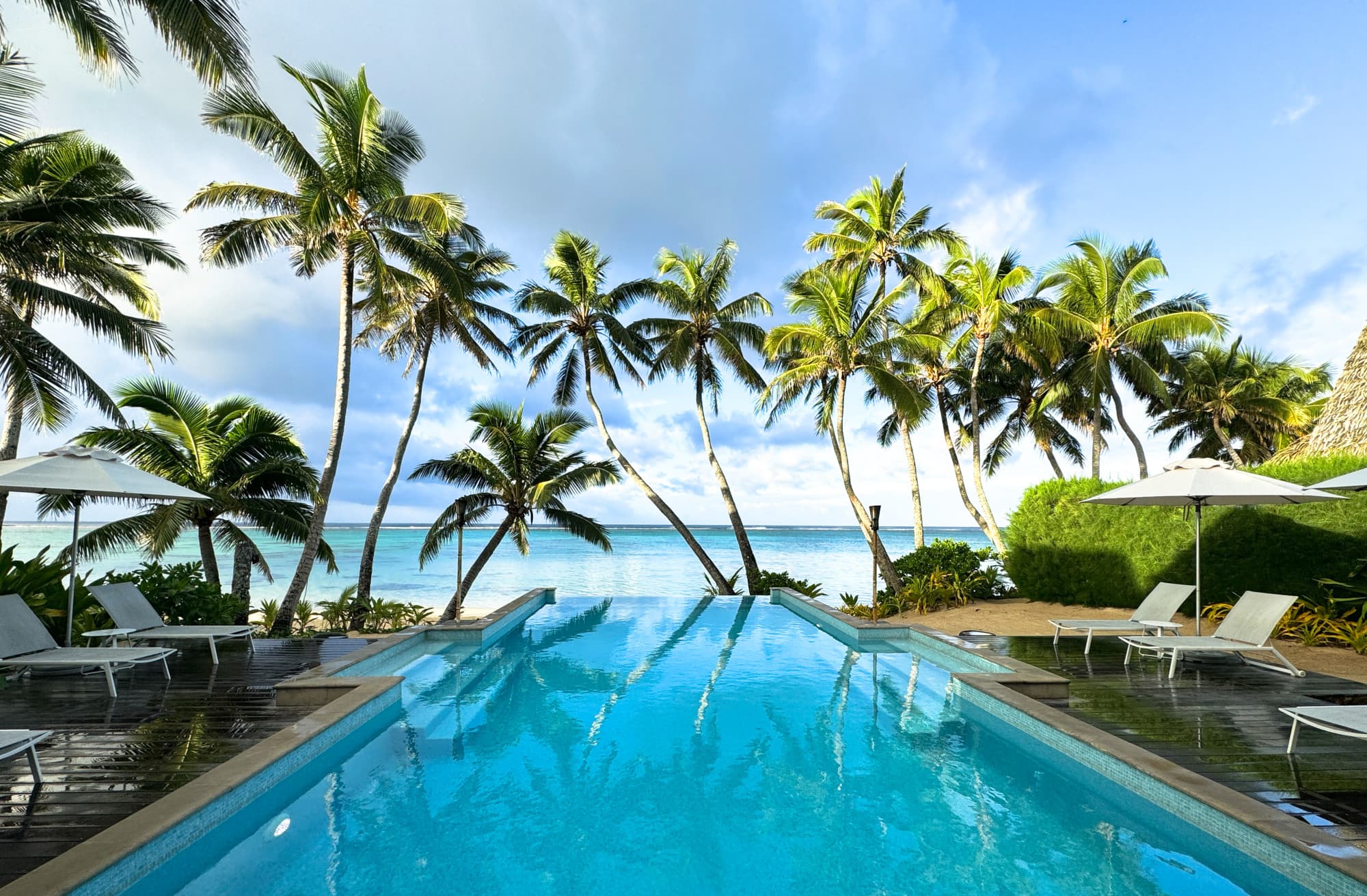 Little Polynesian Resort Rarotonga, pool and lagoon view, with palm trees, sunny skies and blue seas.