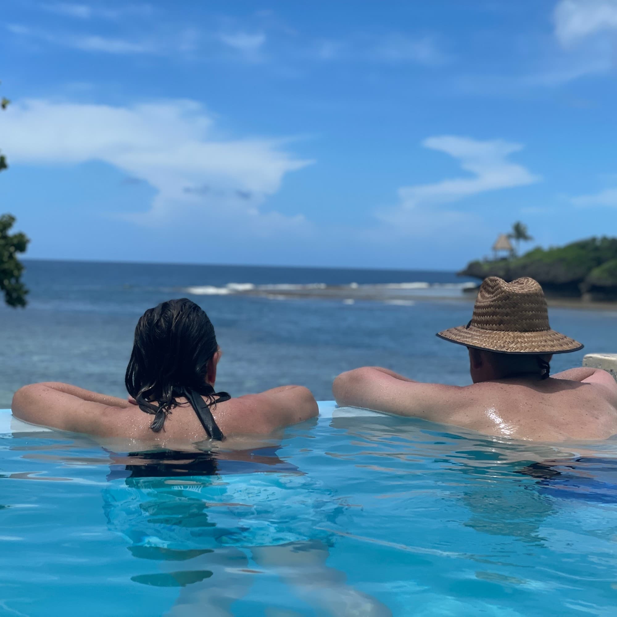 A couple in a swimming pool resting on the edge facing the ocean