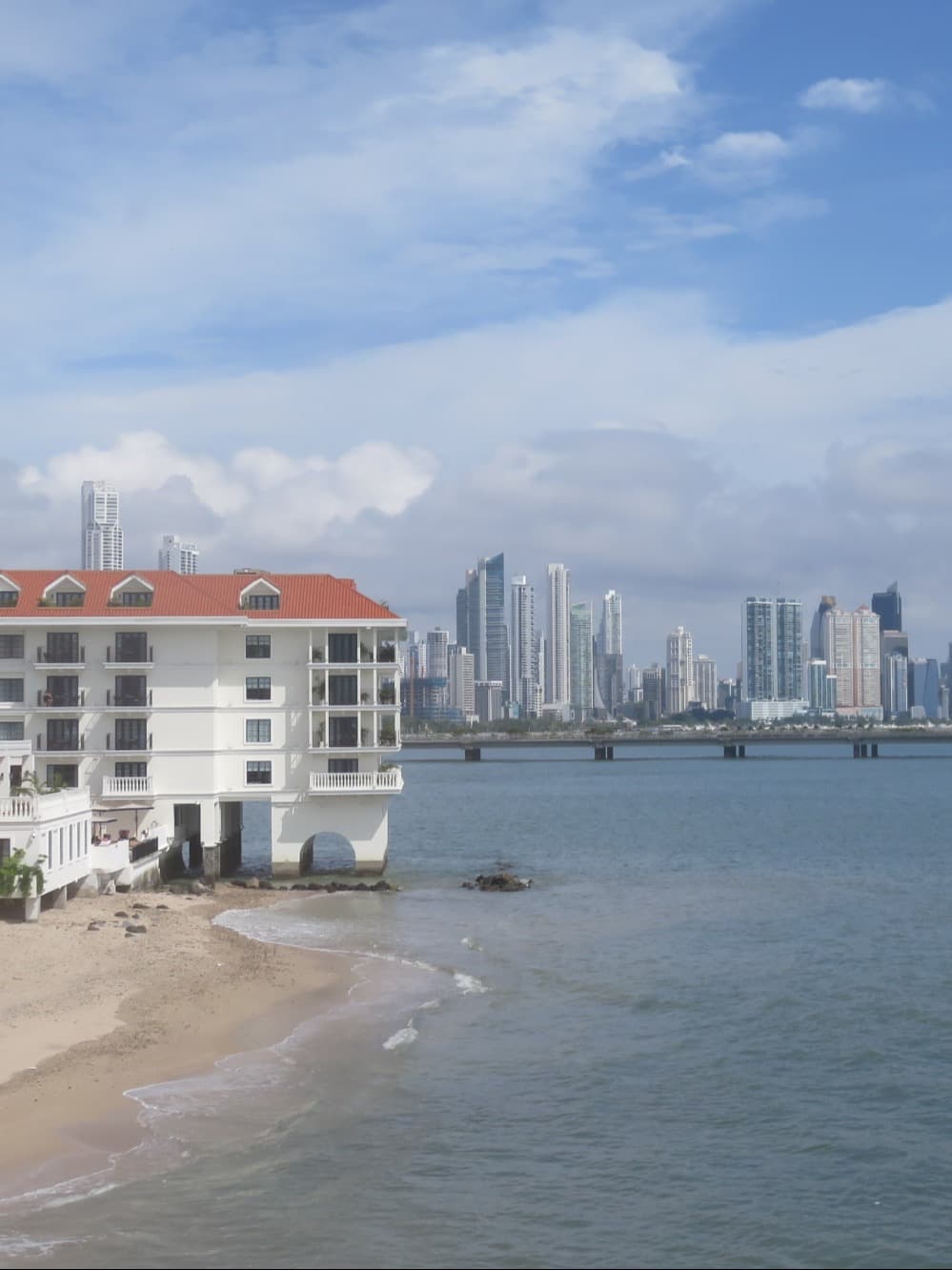 The Sofitel Legend Casco Viejo Panama City from the exterior - a white building on the water with a red roof and the city skyline in the background - Suzanne Tomlin