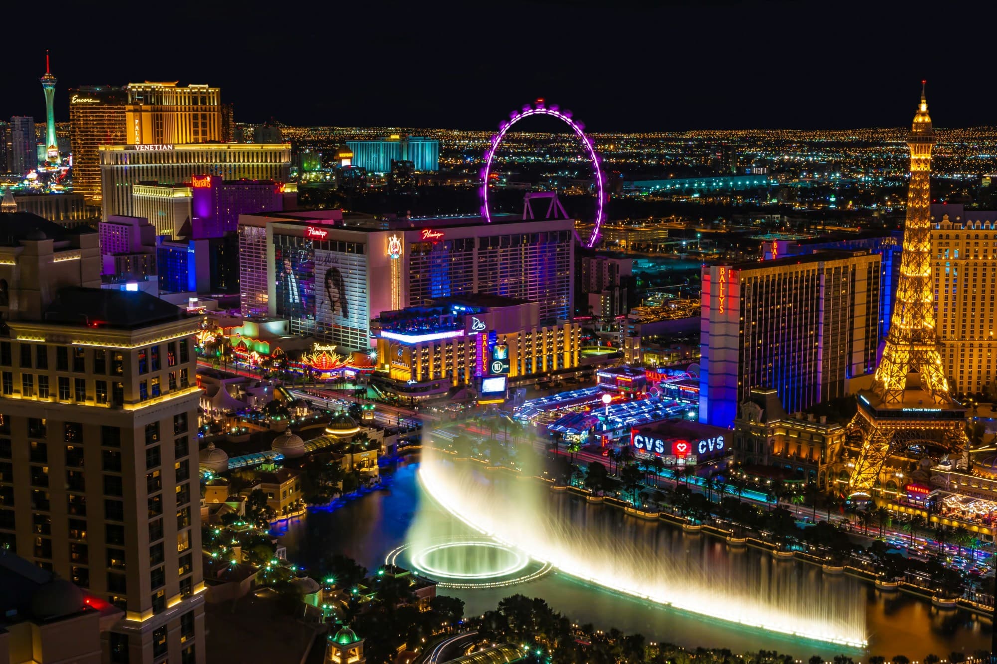 A view of a fountain, a ferris wheel, the mock Eiffel Tower and the Las Vegas strip lit up at night.