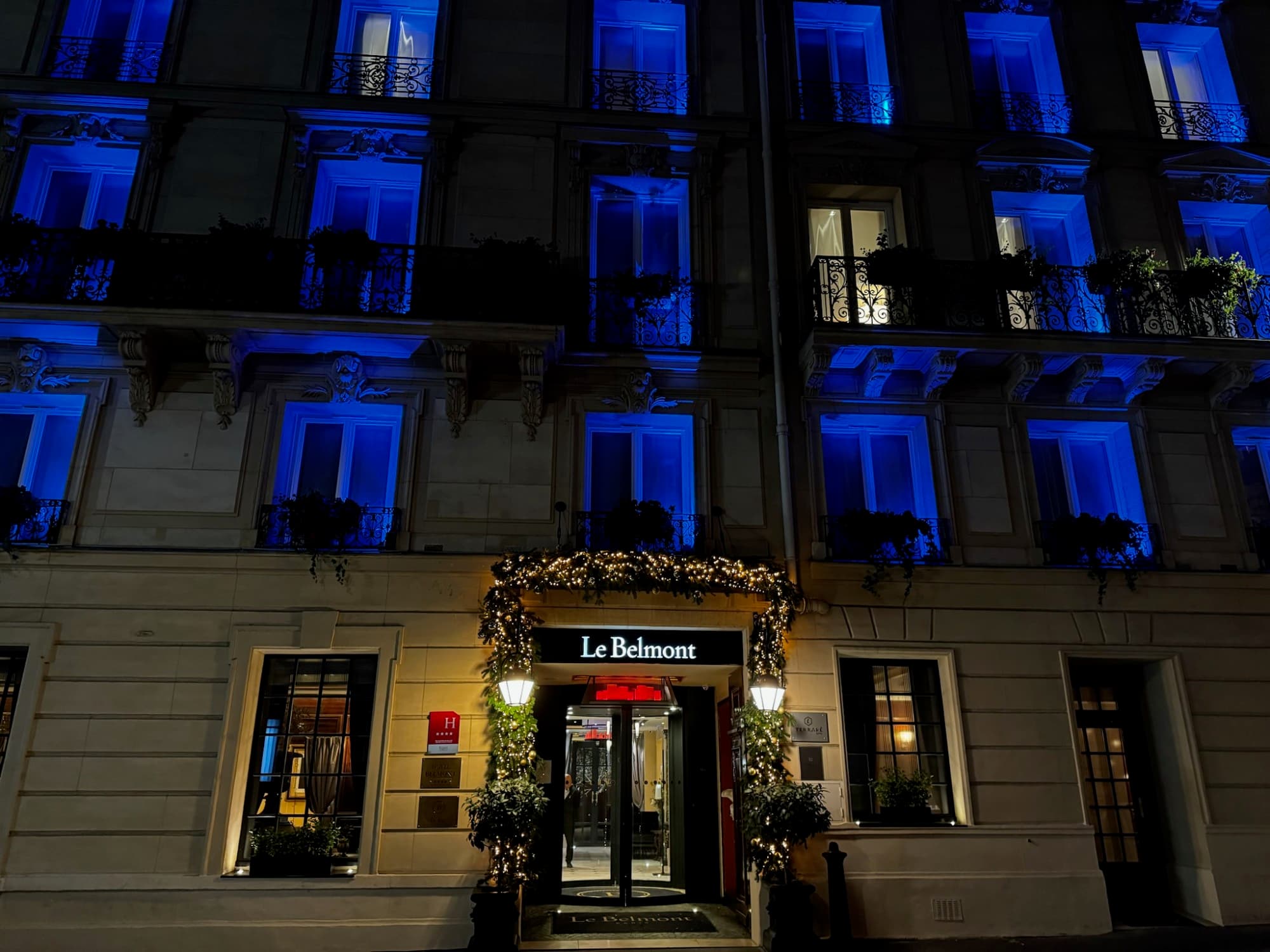 A street view of Le Belmont, with a doorway surrounded by greenery and several floors of blue-lit windows