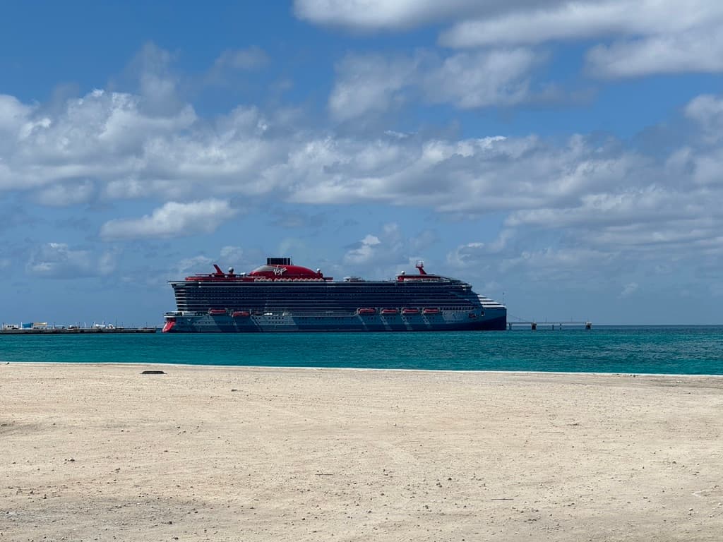 A view from a beach of a large Virgin Voyages cruise ship sailing