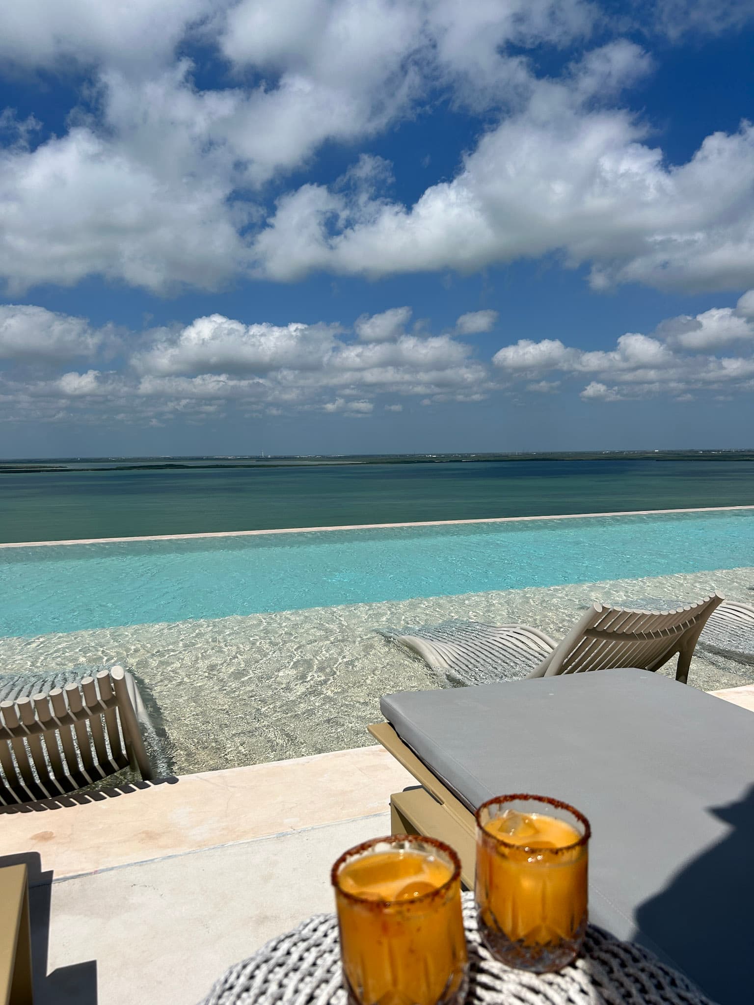 A hotel balcony and infinity pool scene with lounge chairs overlooking the sea and sky on a sunny day.