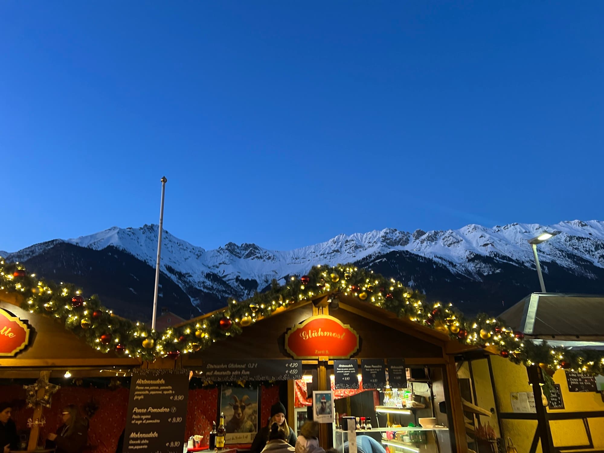 A view of snow covered mountain tops and holiday lights lining the top of a outdoor hut selling goods at a holiday market.
