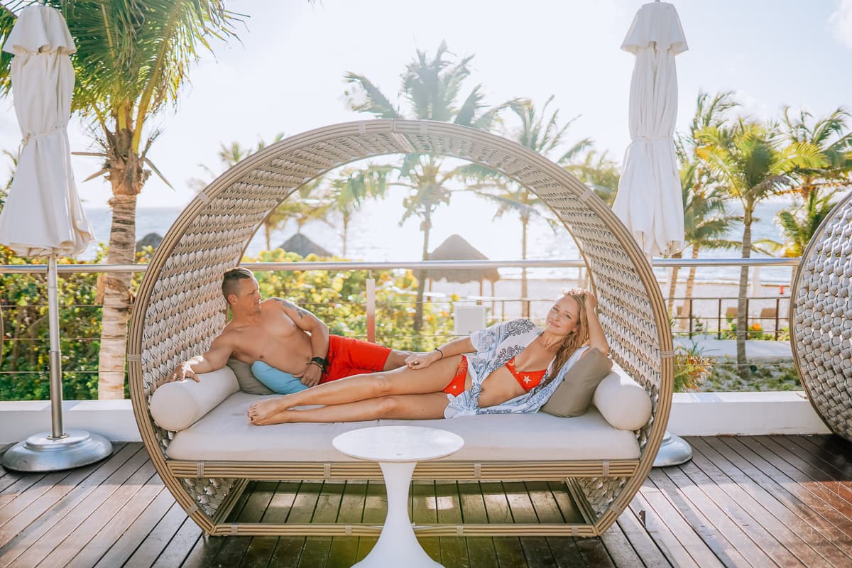 A couple lounging in a cabana on a wooden patio with white umbrellas and palm trees in the surrounding areas.