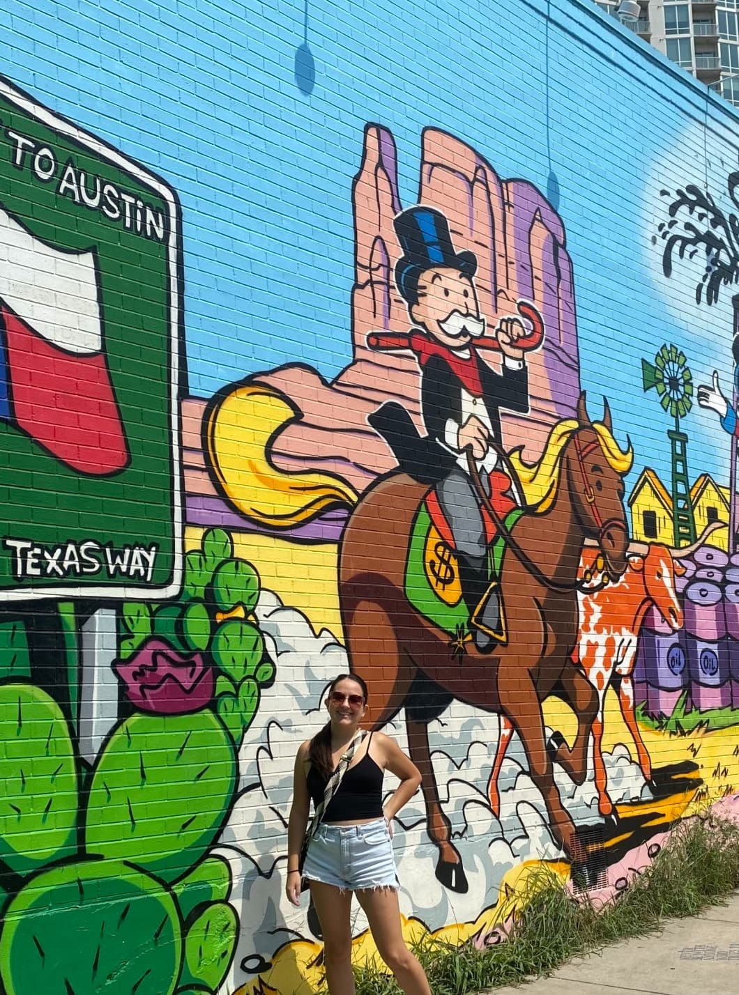 A painted mural with various images including a Texas flag, cactus, horse, people, rocks and more with a woman posing in front of it.