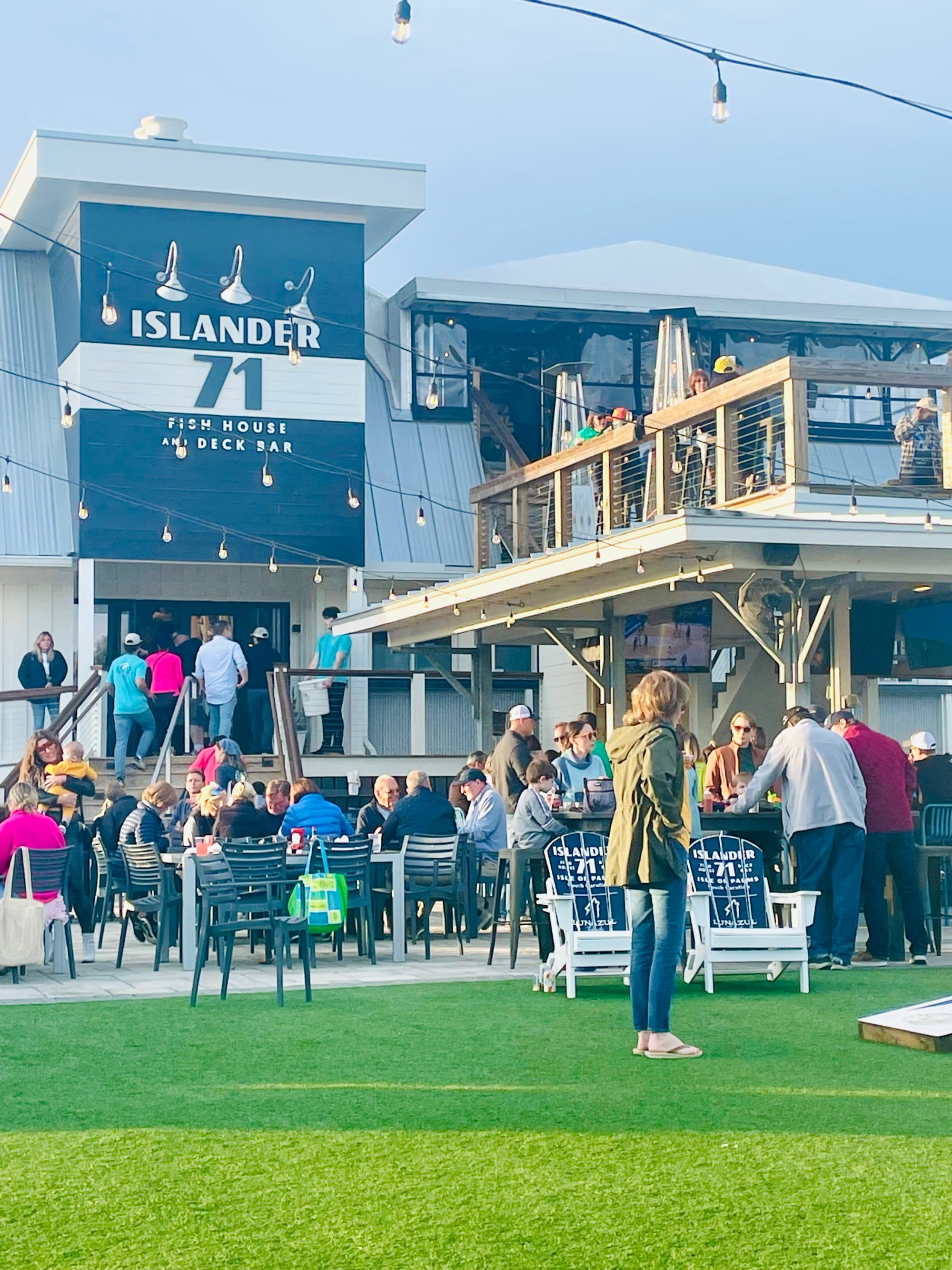 A group of people dining and socializing outside on a patio and lawn. There are string lights, a restaurant exterior and a wooden balcony in the surrounding areas.