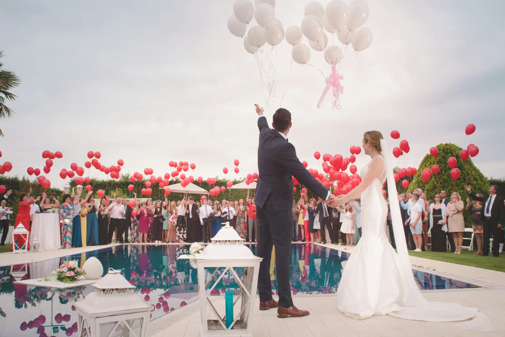 A bride and groom standing next to a pool with wedding guests surrounding and balloons in the air.