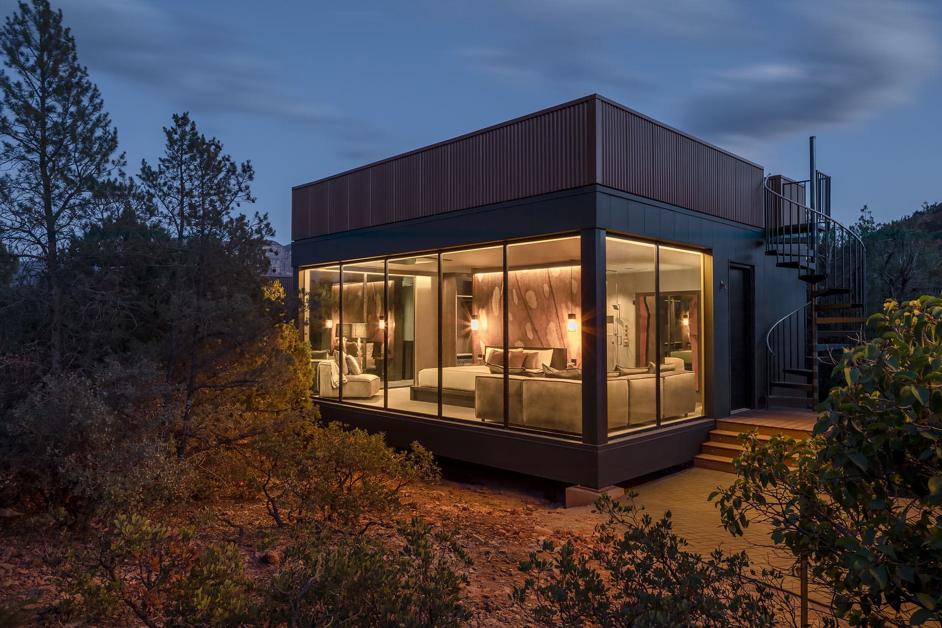 The exterior of a room at Ambiente in the middle of nature at dusk, with floor-to-ceiling windows showcasing a bed and couch inside.