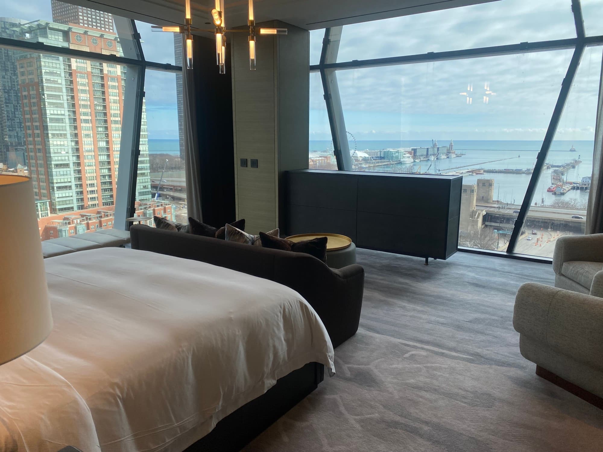 A view of a hotel bedroom with geometric windows, a white bed, modern chandelier lighting, dark wooden credenza and a white chair on the right side.