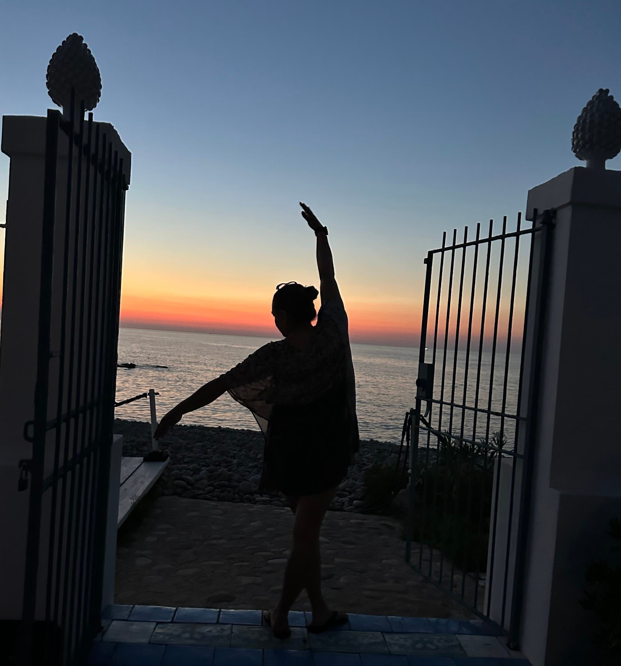 Image of April Bueno posing between two pillars and an open gate at sunset in front of the ocean