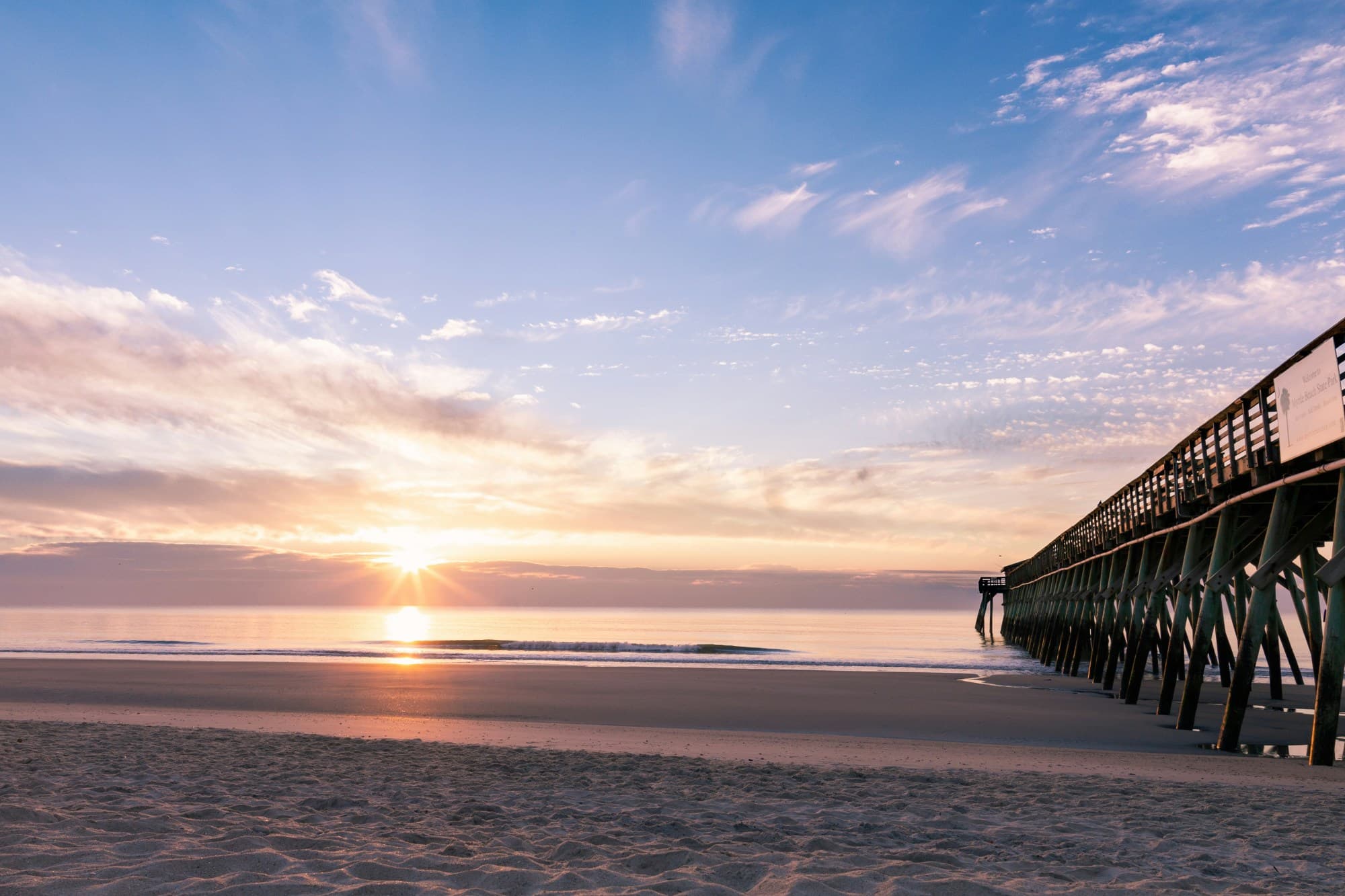 A view of a sandy beach with a wooden boardwalk to the right and the sun setting over the water.
