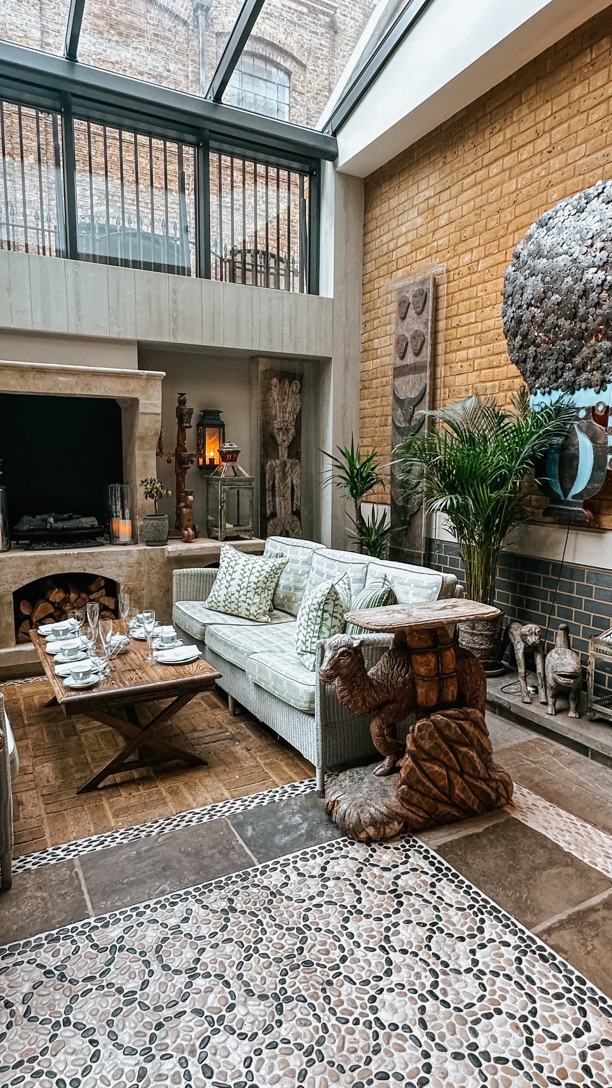 A beautiful hotel lobby with mosaic tile flooring, a white sofa, coffee and side tables, windows, glass ceilings and decorative plants near various pieces of art.
