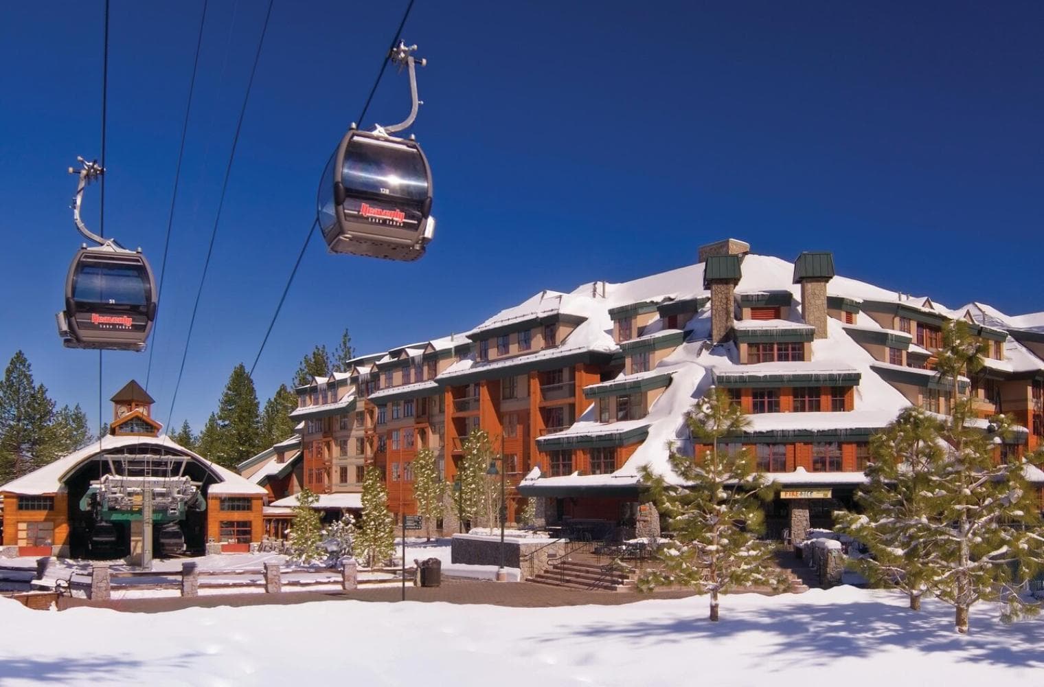 A gondola next a large ski lodge covered in snow and surrounded by pine trees under a clear blue sky.