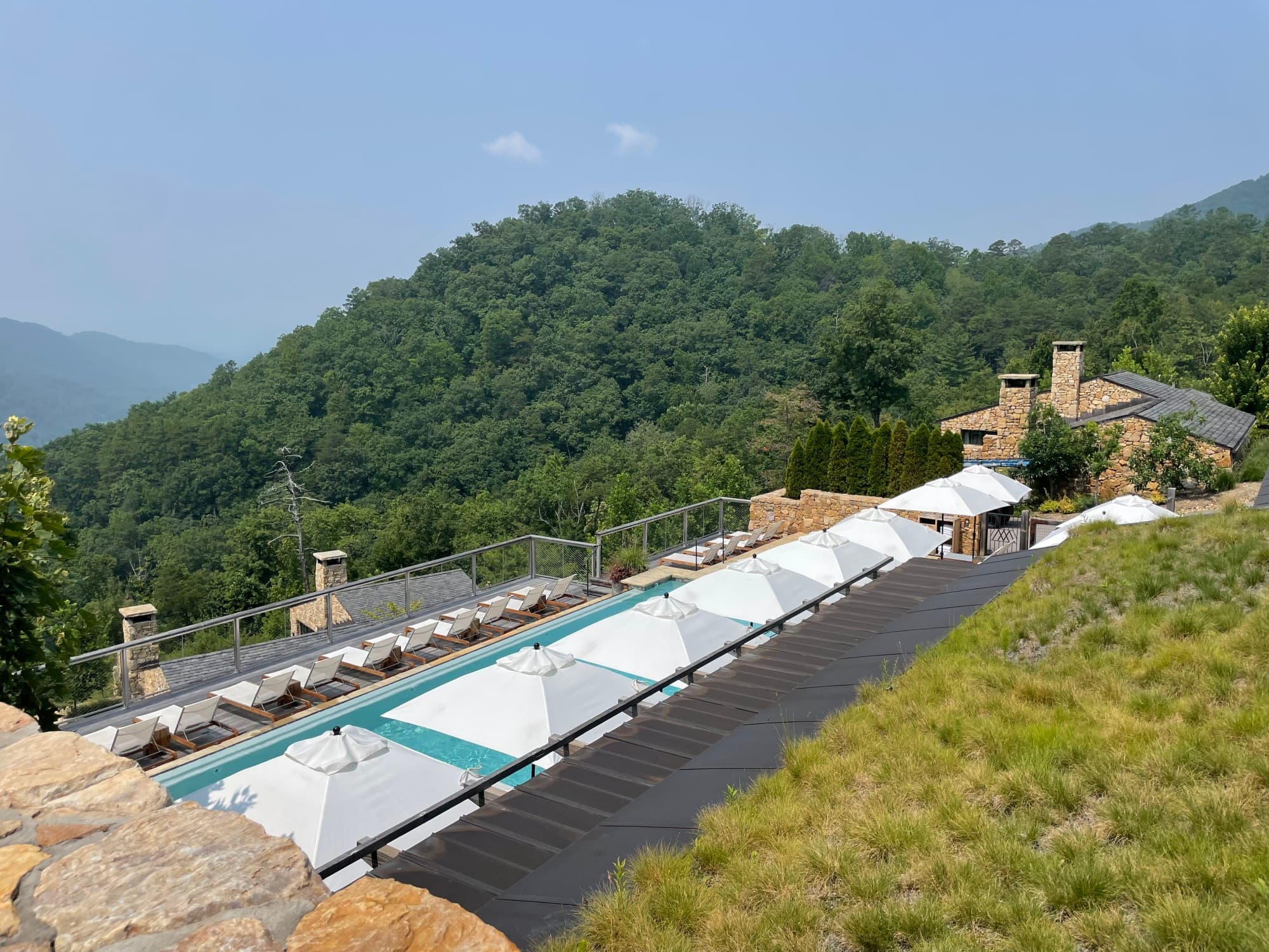 A top down view of a swimming pool deck complete with rows of white umbrellas and lounge chairs overlooking a lush, green mountain.