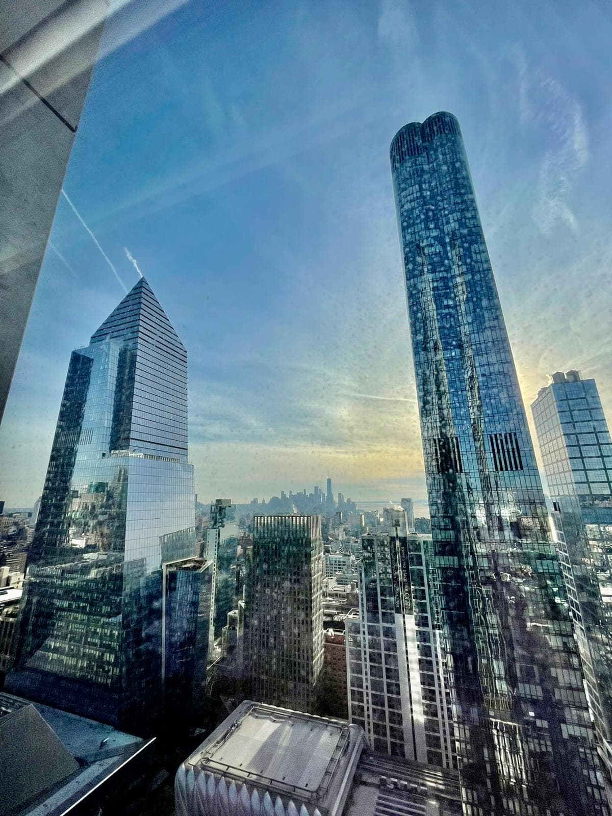 A view of the reflective skyscrapers in New York City with the sun peaking out of the clouds in the distance.