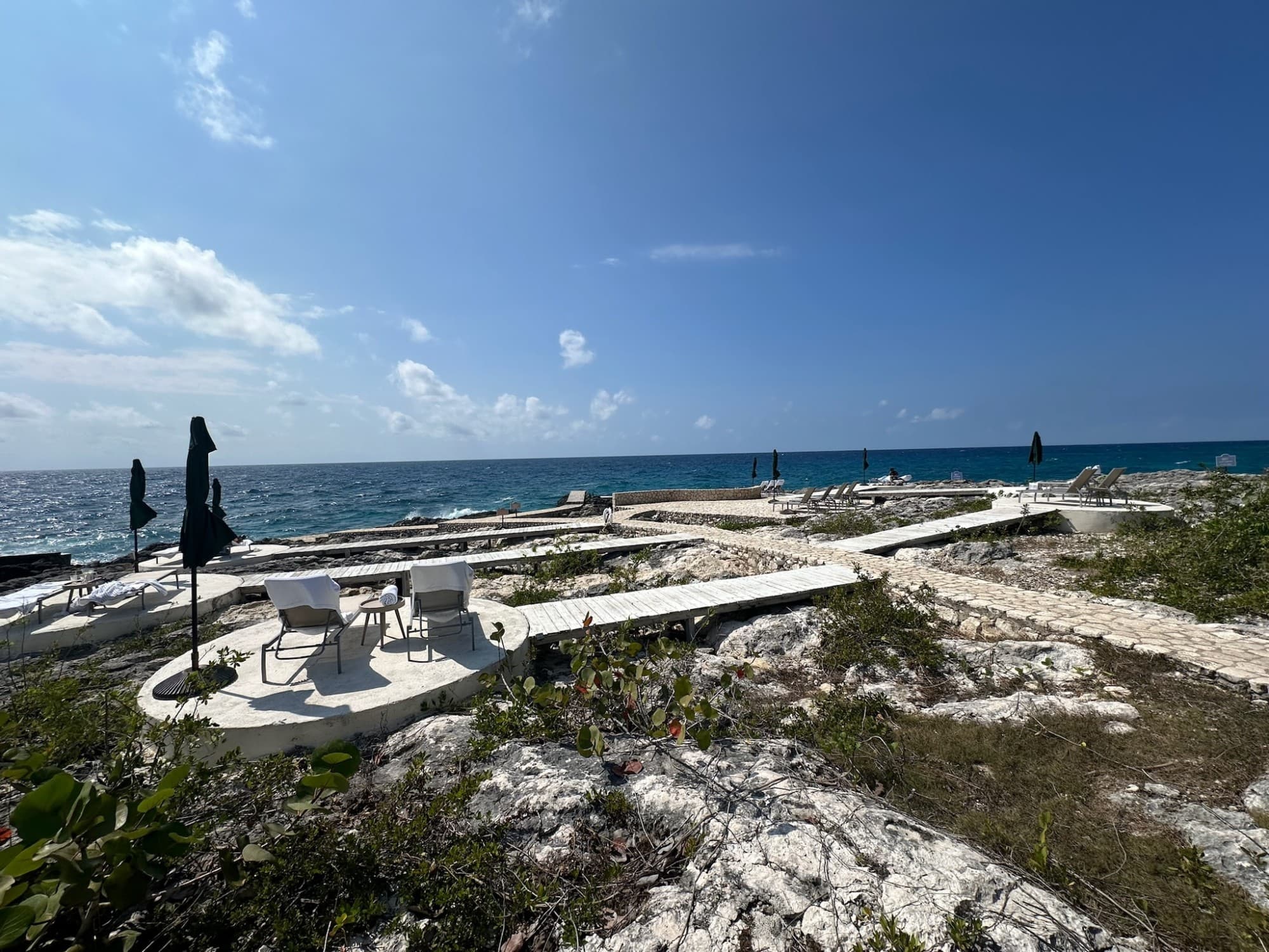 A beautiful view of a sandy beach with white boardwalks leading to a seating area complete with two lawn chairs and an umbrella overlooking the blue water.