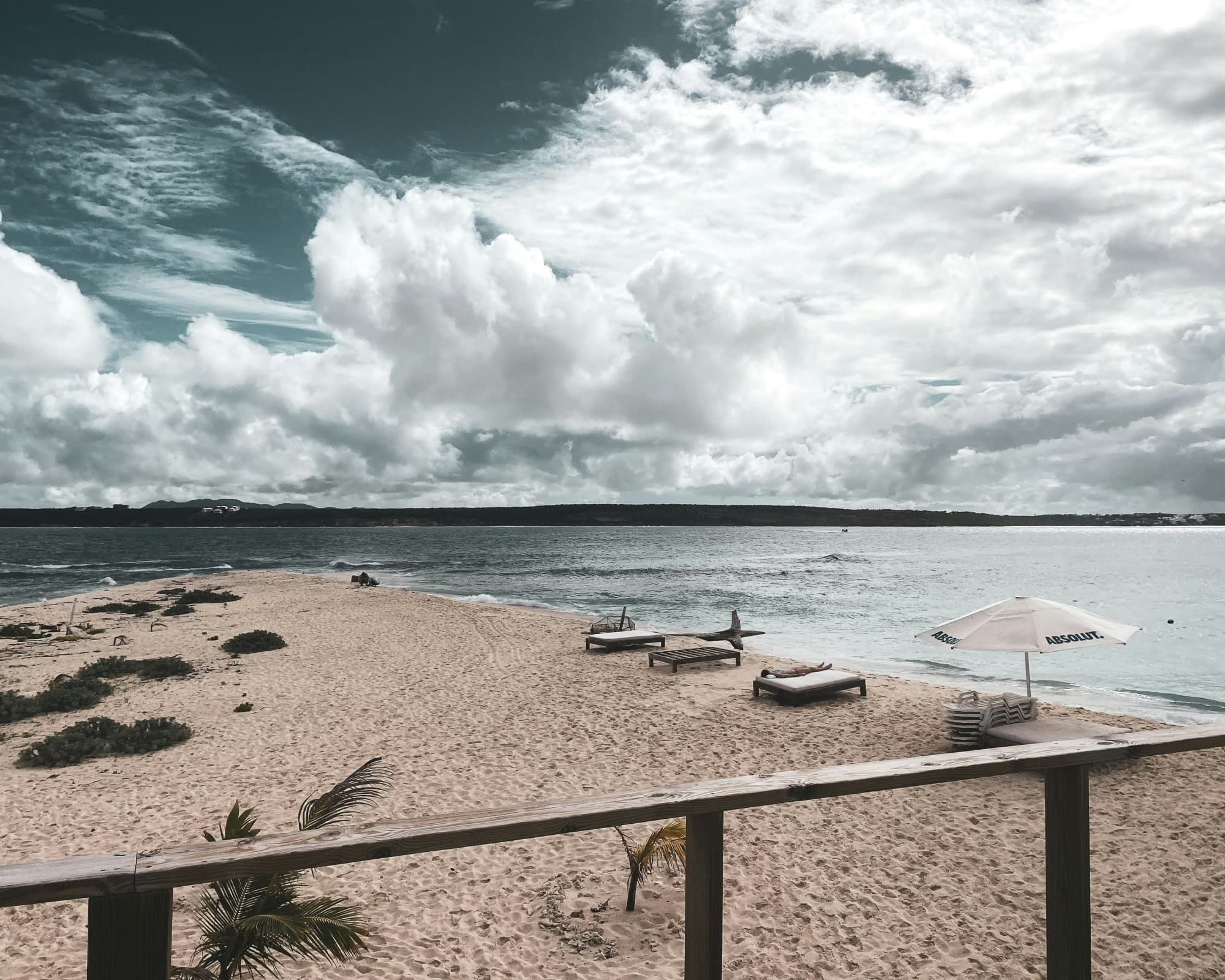 A filtered picture of a wooden railing with a sandy beach in the background against the sea. There are vacant lawn chairs and one white umbrella on the beach, all situated under a vast, cloudy blue sky.
