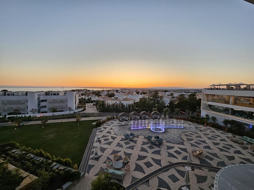 An aerial view of a terrace surrounded by white buildings, grass, a lit up fountain feature and a golden sunset in the background.