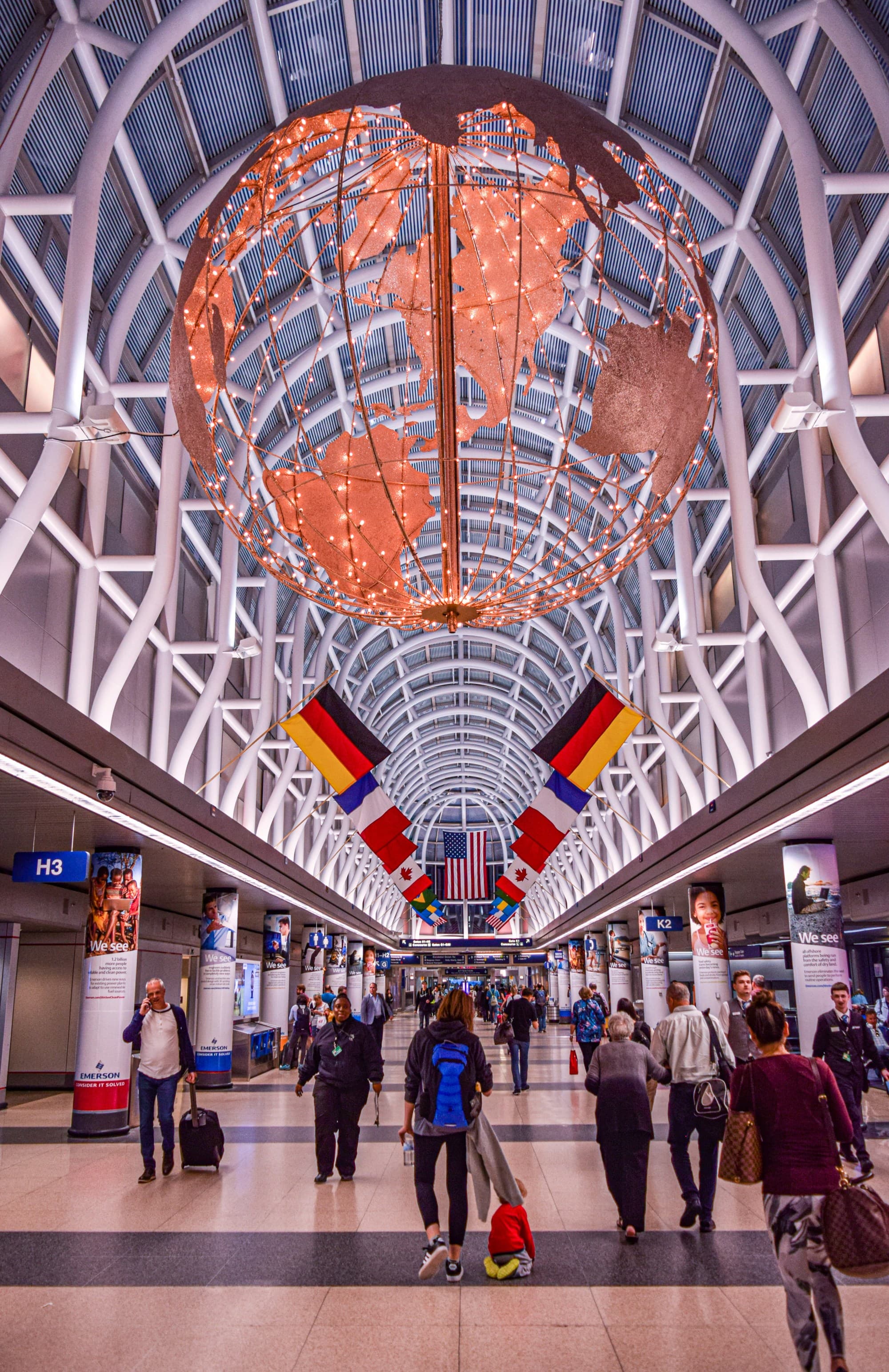 An interior view of an airport with a large metal globe on the ceiling surrounded by white beams and people walking around beneath various flags.