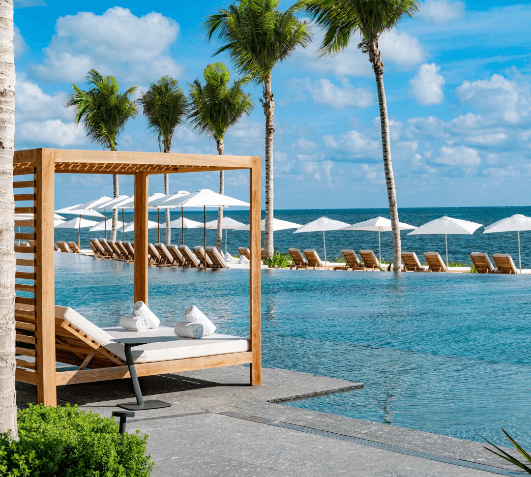 A swimming pool with wooden cabanas, white umbrellas and palm trees.