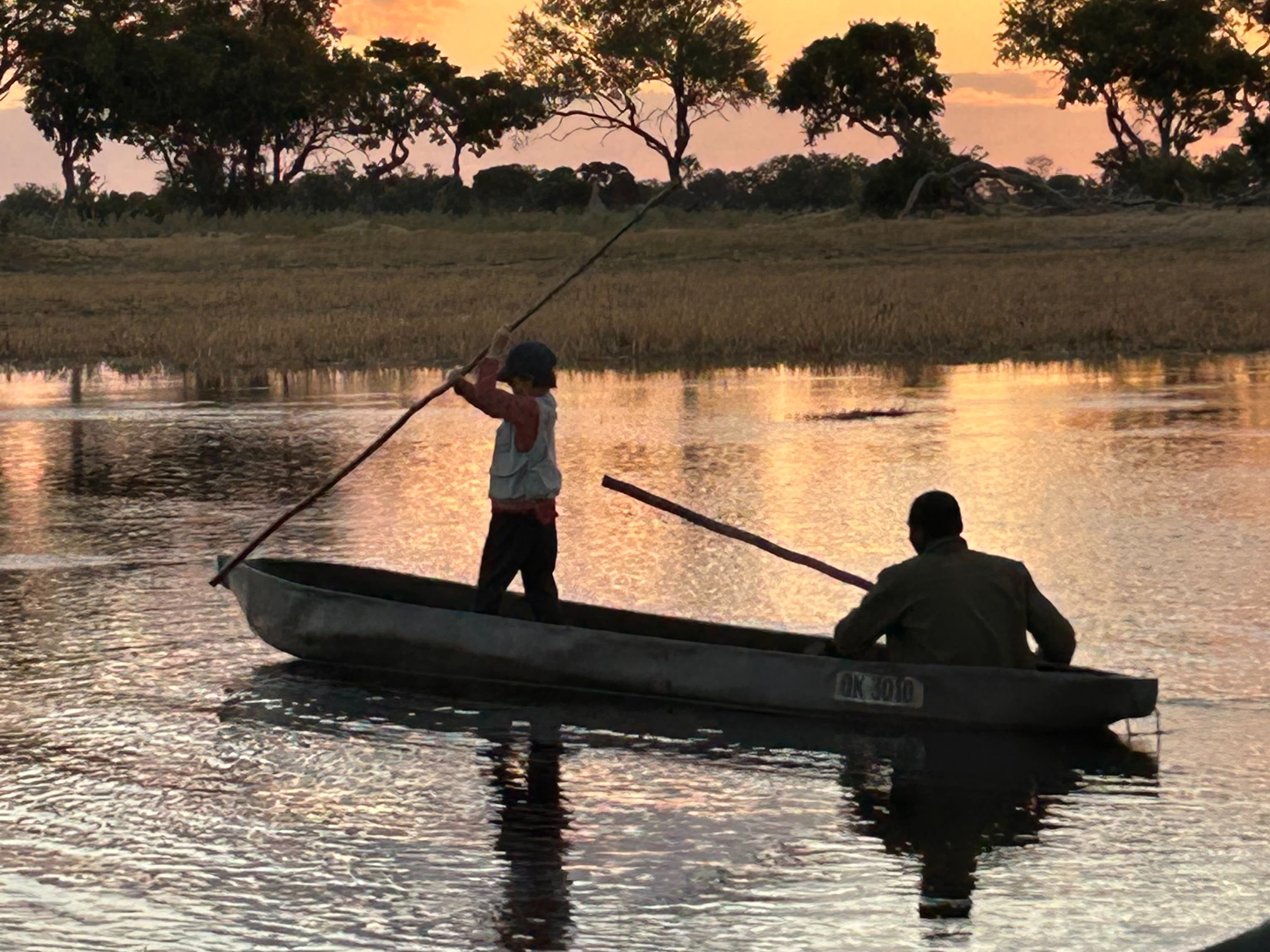 Two individuals fishing on a boat during a sunset with trees in the background.