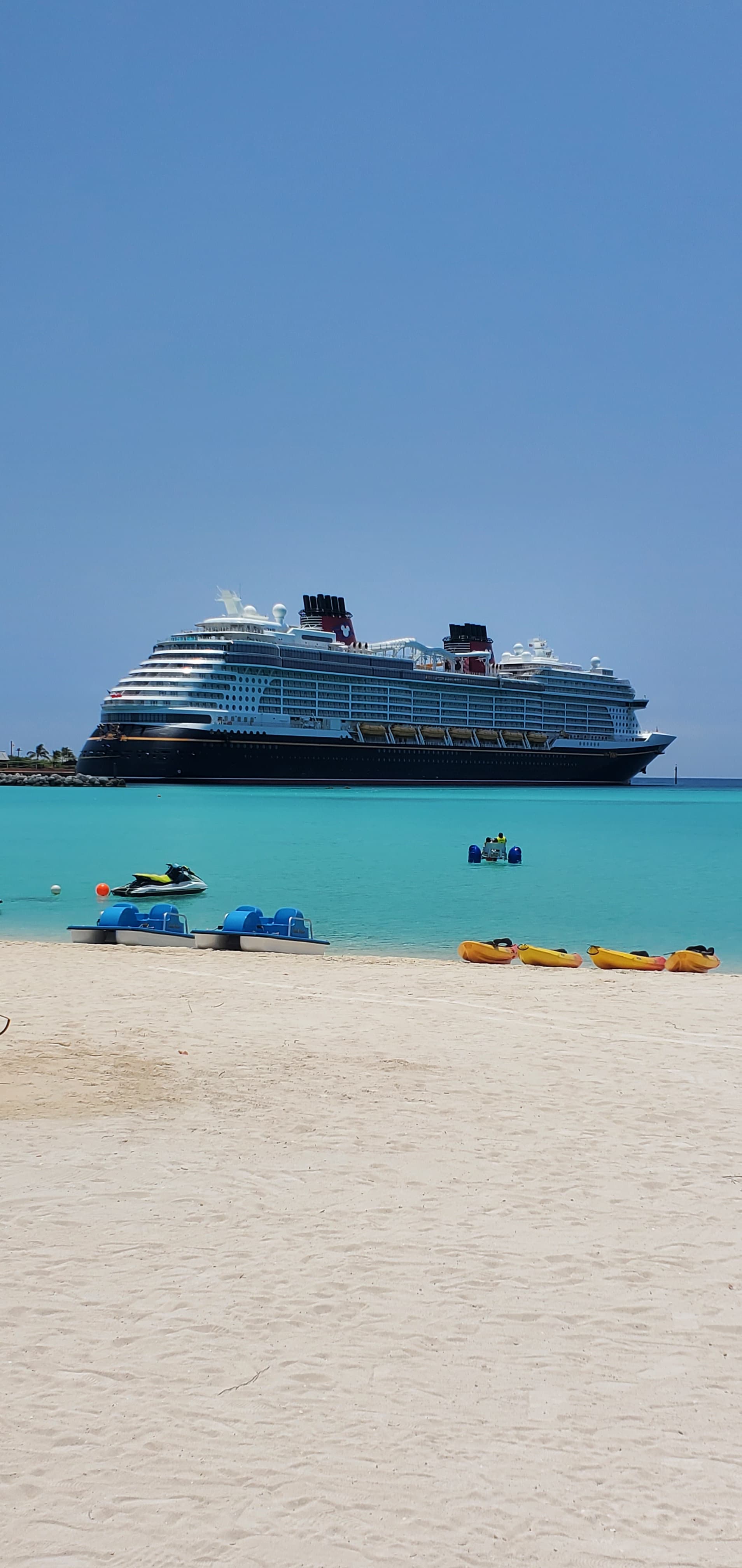 A cruise ship docks in the harbor across from a sandy beach on a clear day.