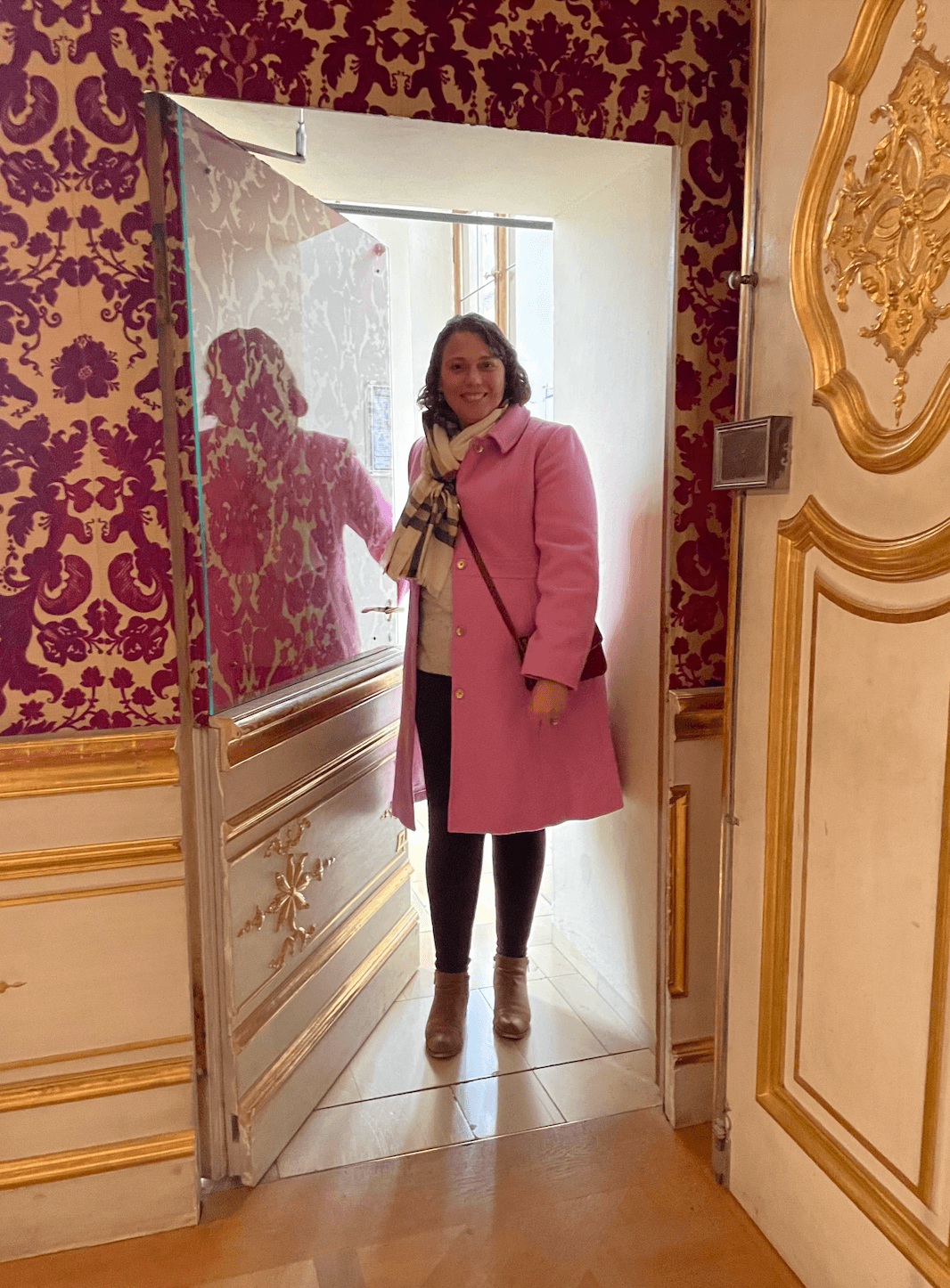 A woman in a pink blazer, white top and black pants posing in a doorway with luxurious wallpaper and gold paneled walls in the surrounding area.
