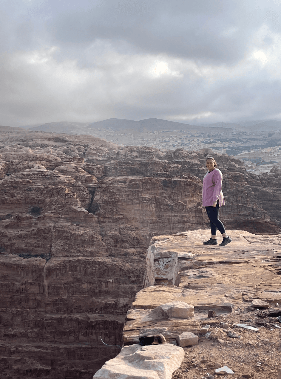 Kathryn wearing a pink top and black pants while standing on the edge of a rocky cliff with large rock formations in the background.