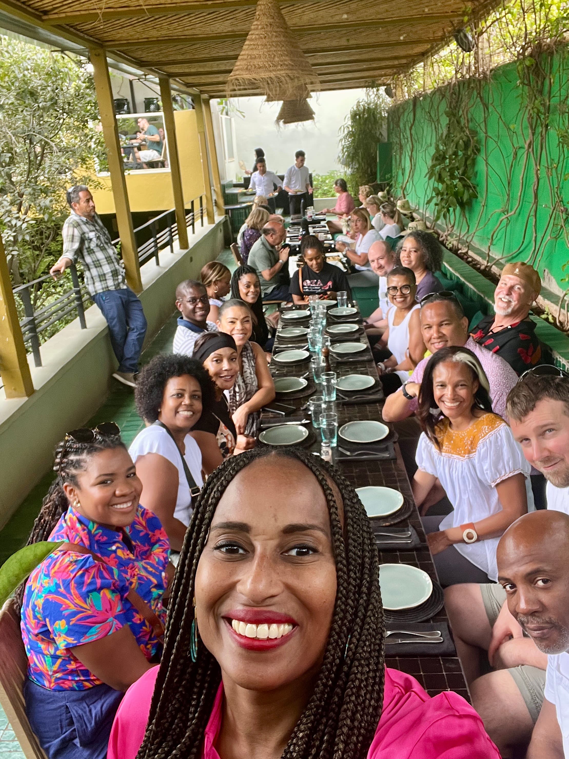 Nadeen posing for a selfie with other travelers seated at a dining table in the background, smiling as well.