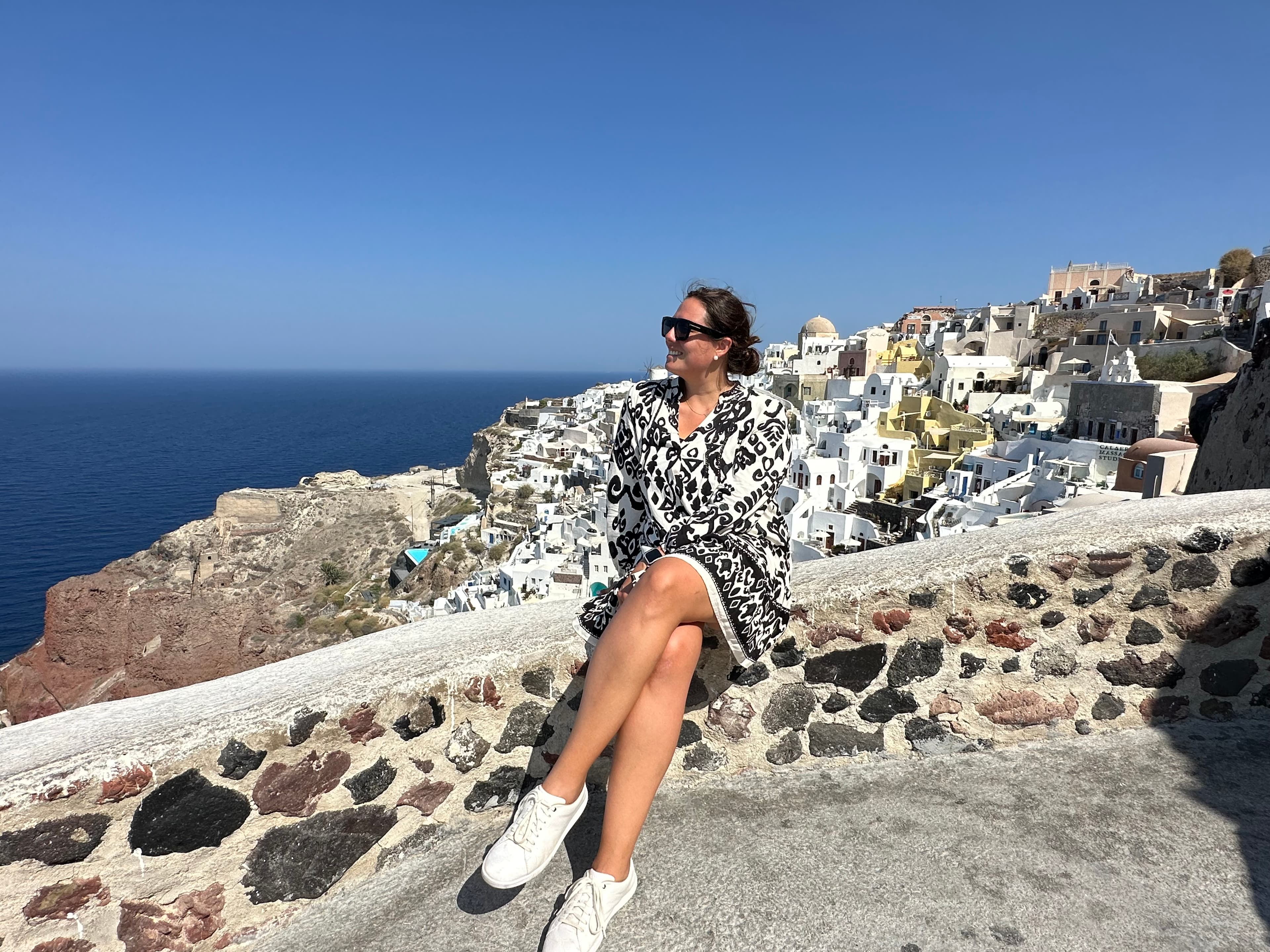 Travel advisor Andrea sitting on a stone wall on a hillside overlooking a beautiful view of buildings and ocean in Santorini