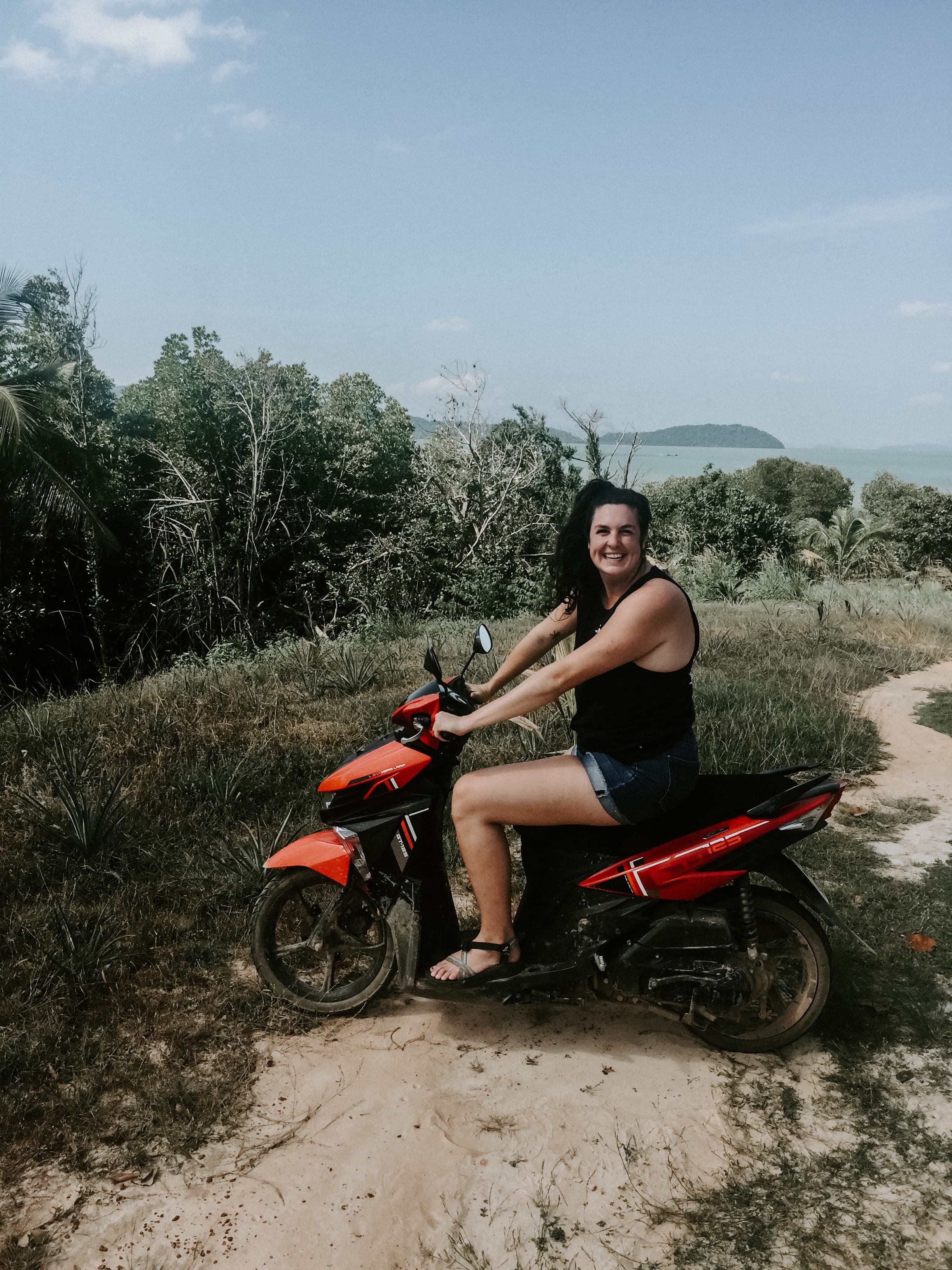 Travel advisor Rachel in a black tank top and shorts riding a red motorbike along a trail with the ocean in view in the distance