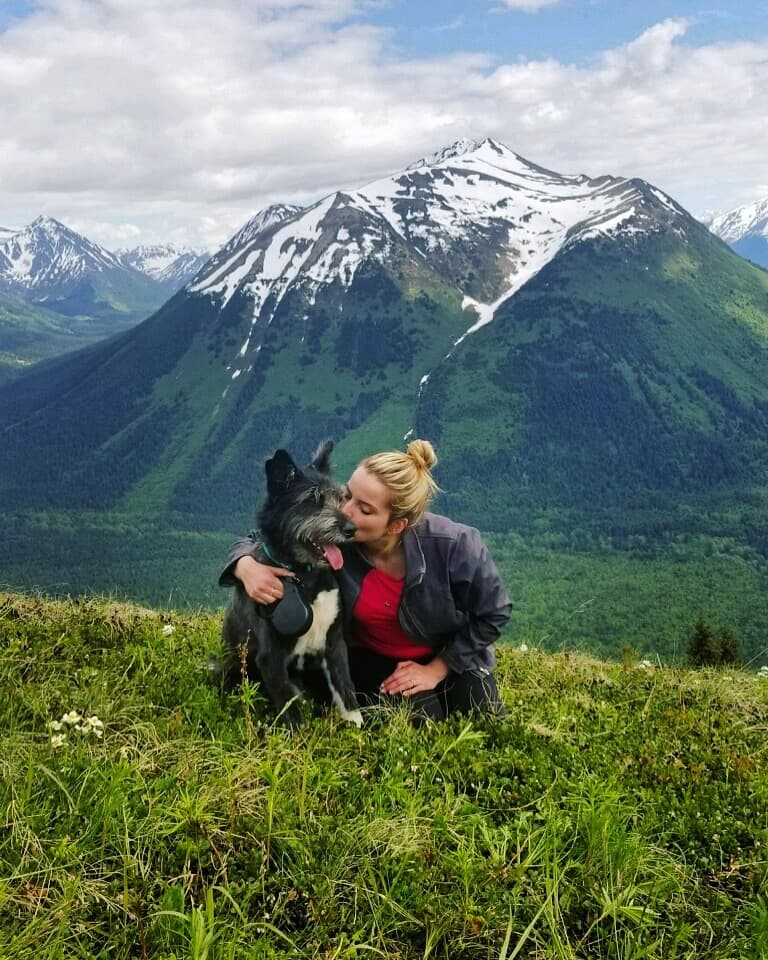 Travel advisor Elizabeth kissing a black and white dog on a grassy field in front of snow-capped mountains