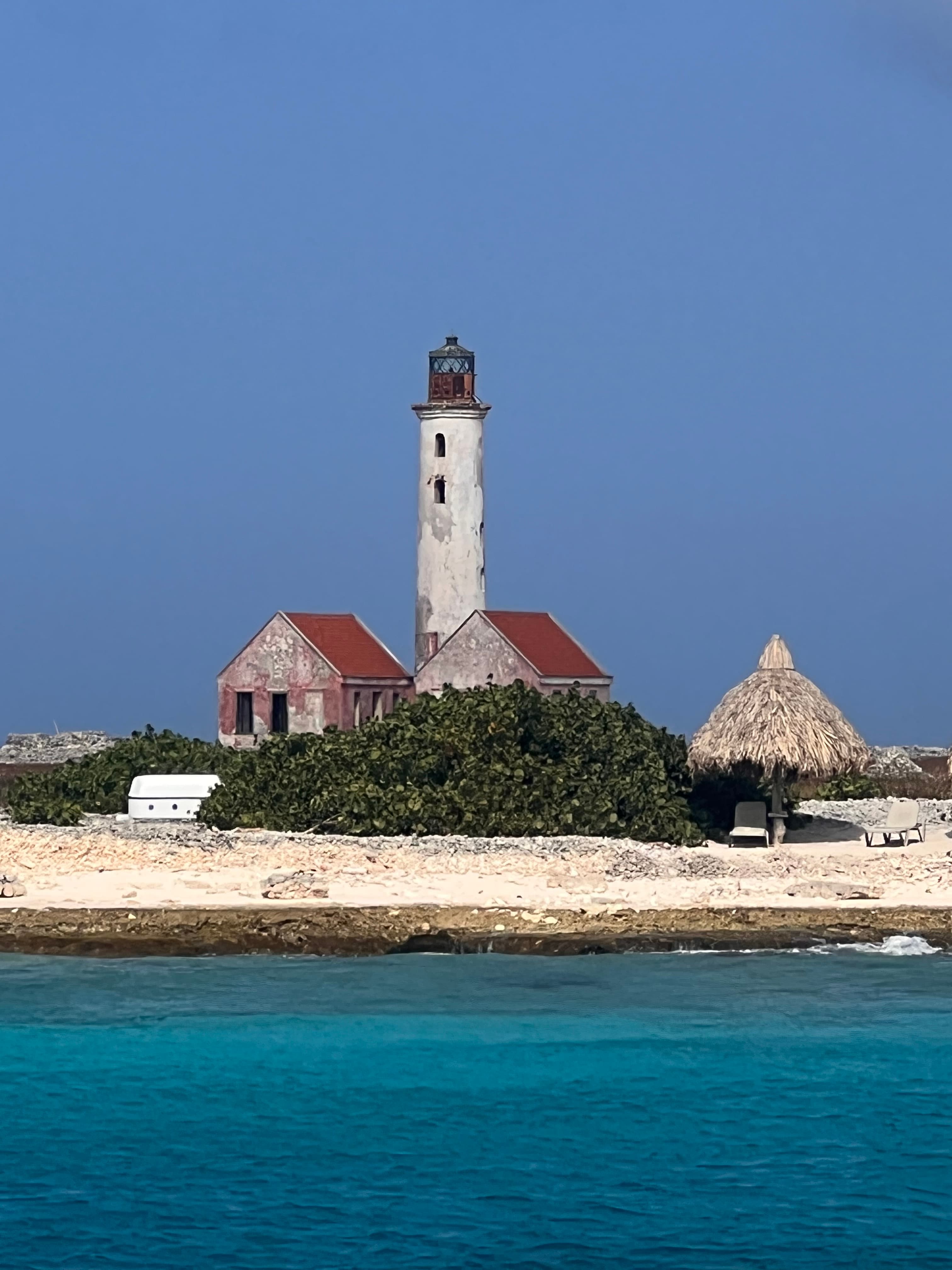 Beautiful view of white lighthouse with two small buildings below with red roofs and a thatched umbella on a small beach.