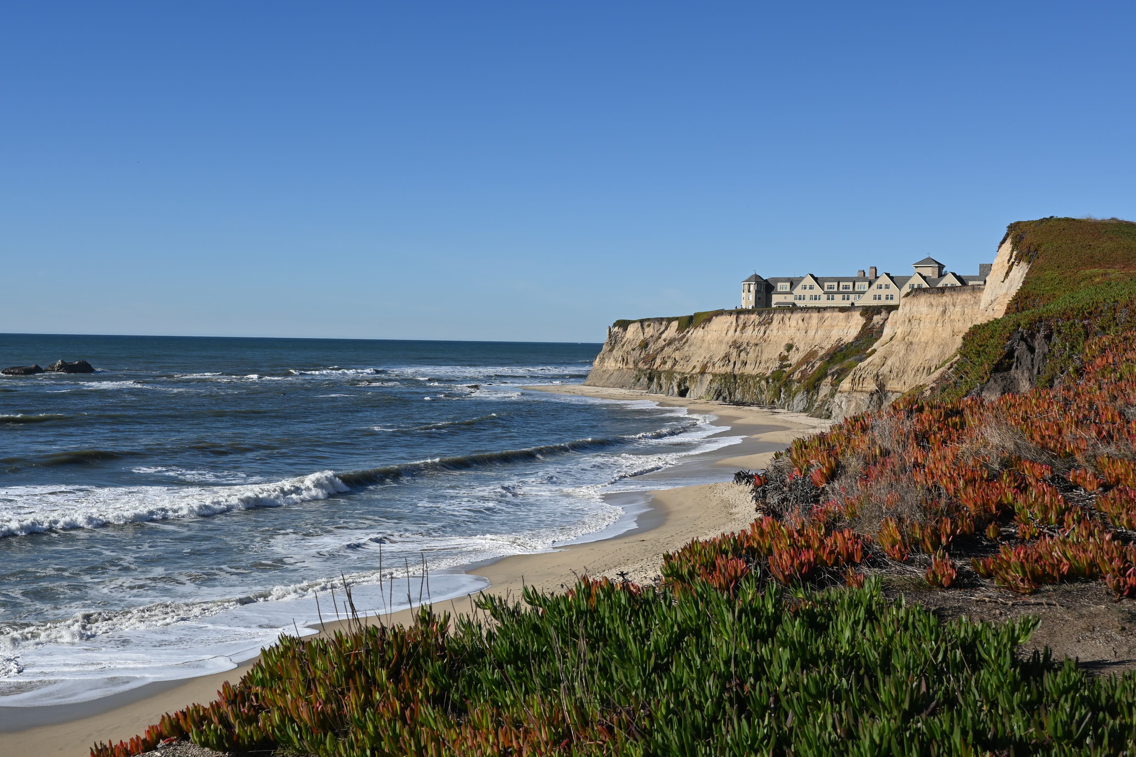 A coastal view of sand-colored cliffs and a beautiful beach with green and orange shrubs around